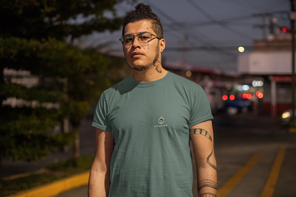 Rain Check Garment-Dyed Tee in Blue Spruce, worn by a man on a street at dusk, with the Rain Check logo and portage.camp on the chest. Digital product mockup.