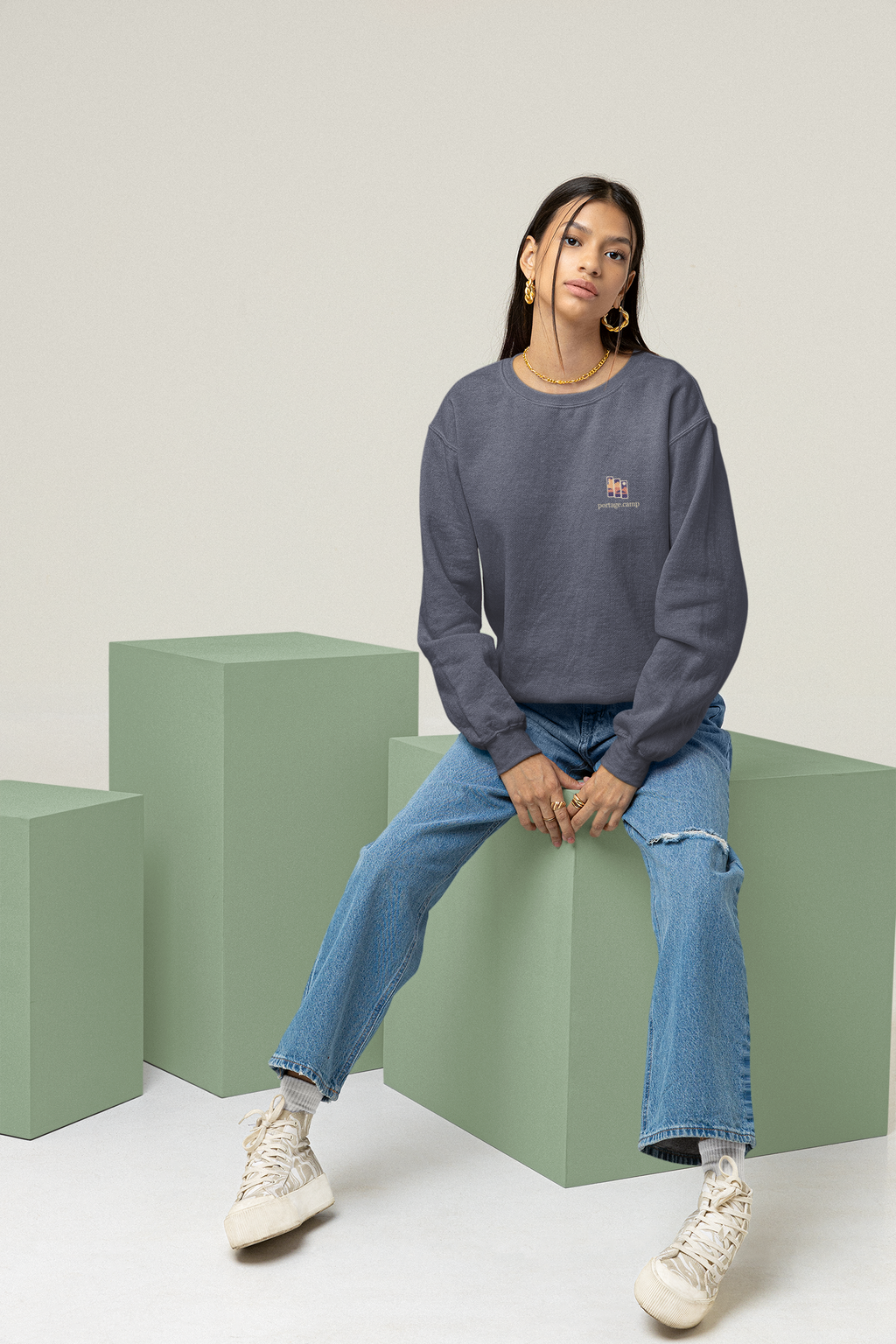 Stay for the Sky Garment-Dyed Crewneck in Denim, worn by a woman seated on green studio blocks, with the Stay for the Sky logo and portage.camp on the chest. Digital product mockup.