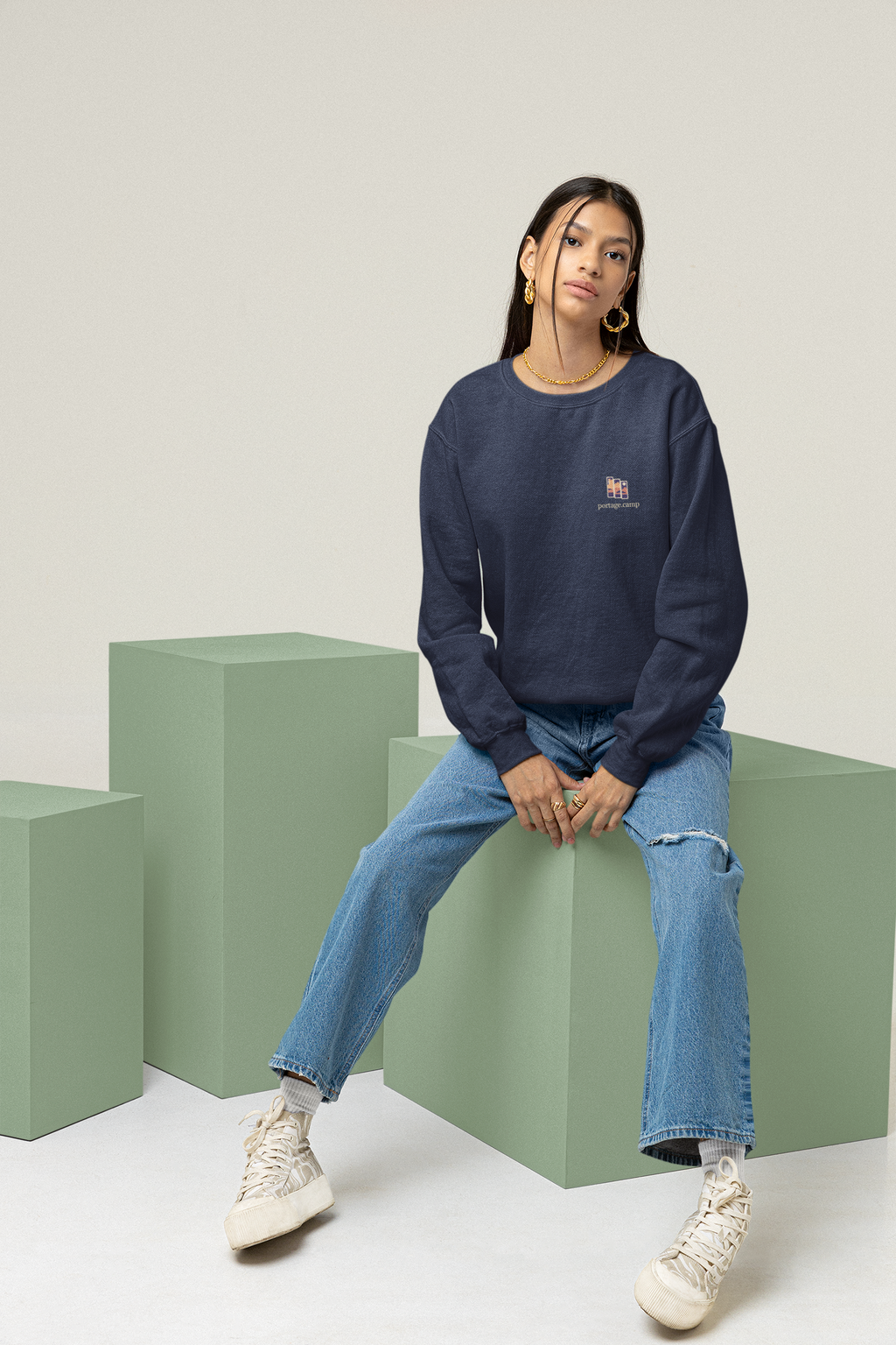 Stay for the Sky Garment-Dyed Crewneck in True Navy, worn by a woman seated on green studio blocks, with the Stay for the Sky logo and portage.camp on the chest. Digital product mockup.