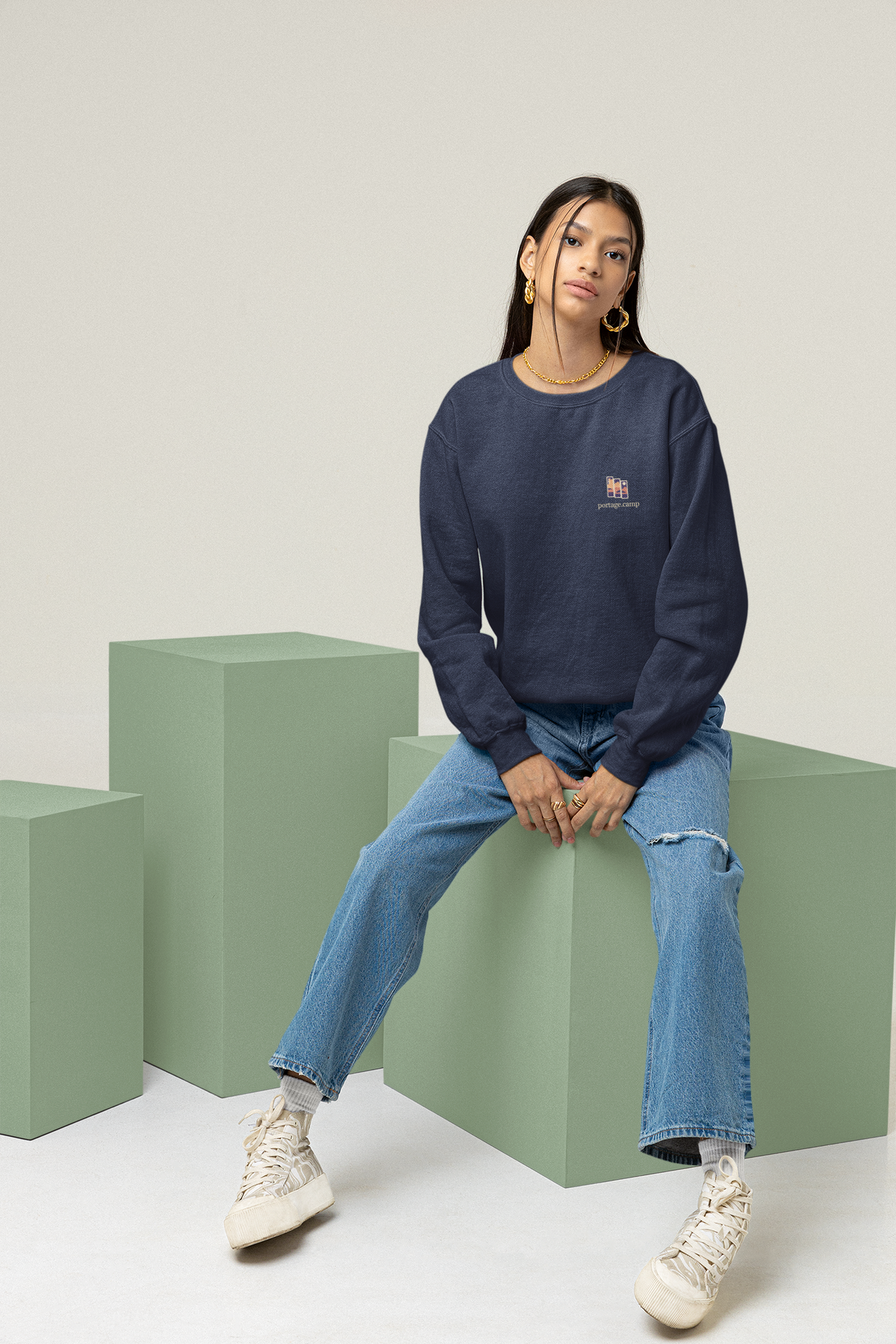 Stay for the Sky Garment-Dyed Crewneck in True Navy, worn by a woman seated on green studio blocks, with the Stay for the Sky logo and portage.camp on the chest. Digital product mockup.