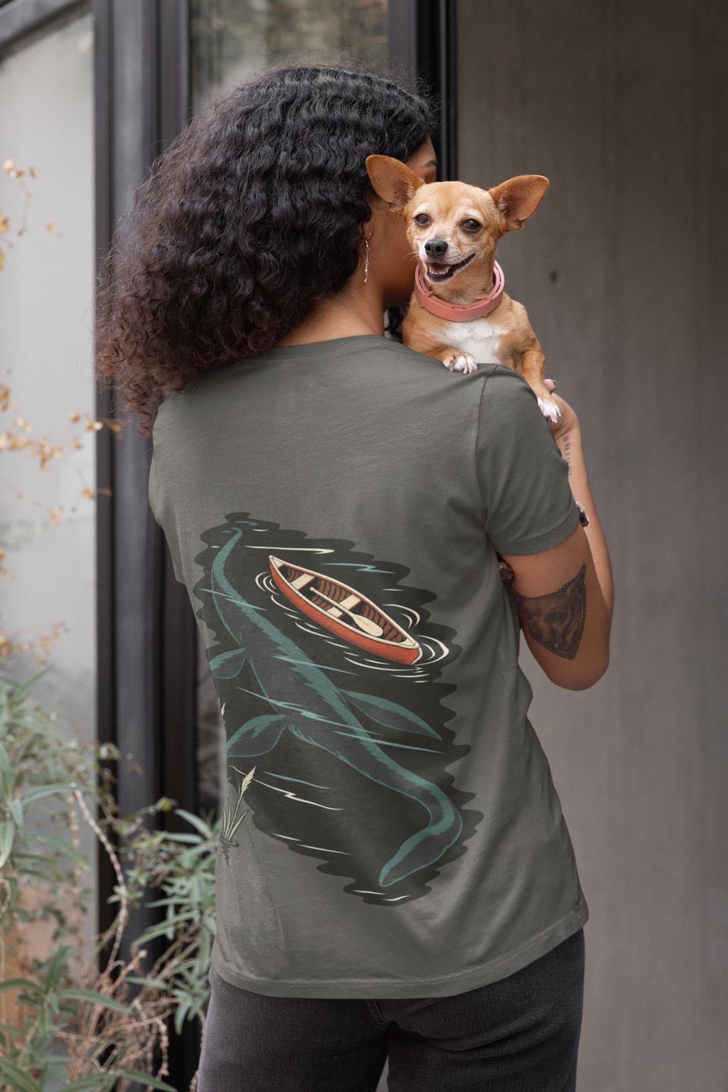 Seen Briefly Garment-Dyed Tee in Pepper, worn by a woman seen from behind holding a small dog, showing the large print on the back. Digital product mockup.