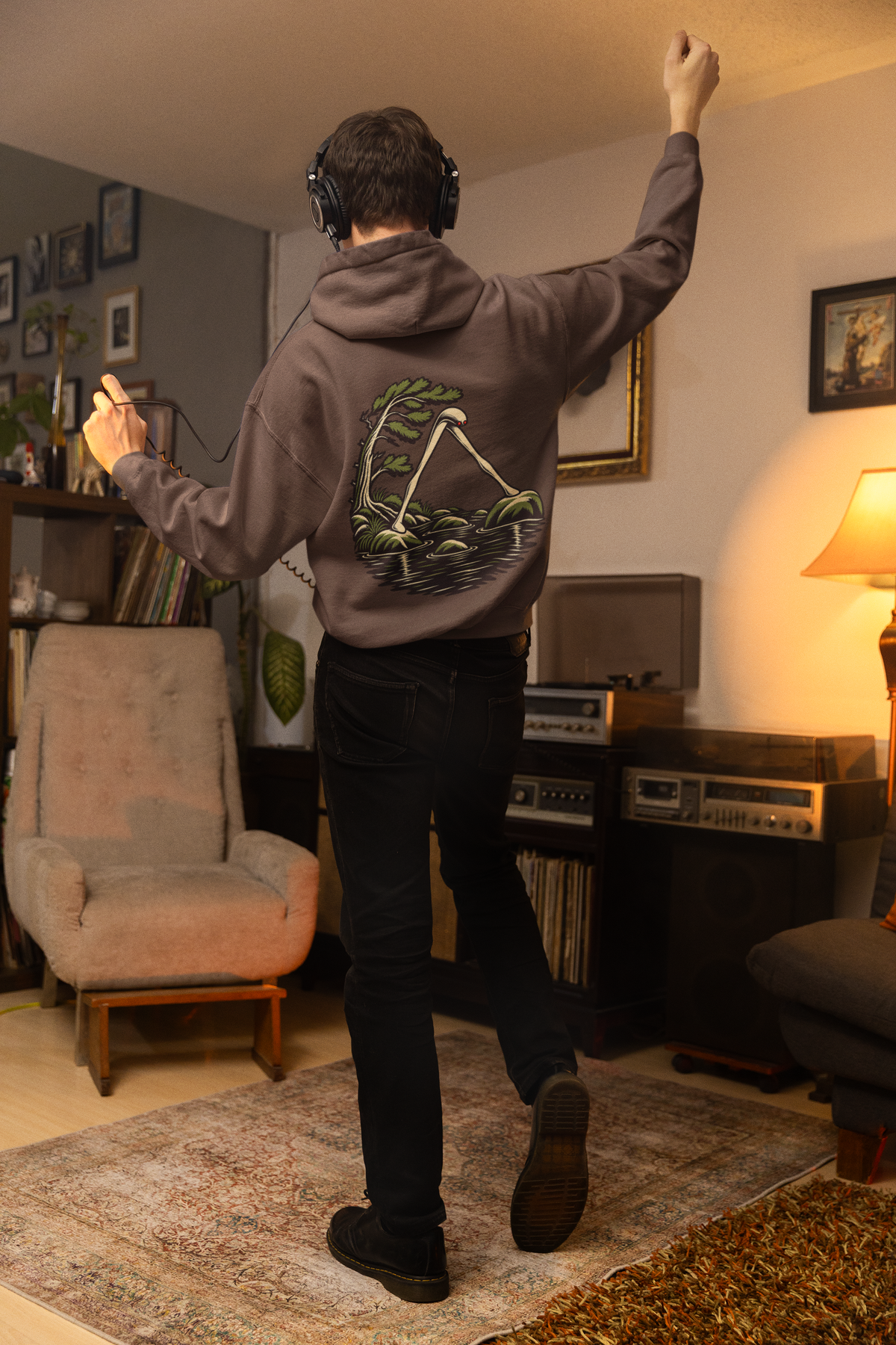 Small Hours Heavyweight Hoodie in Brown, worn by a man seen from behind in a living room at night, showing the large print on the back. Digital product mockup.