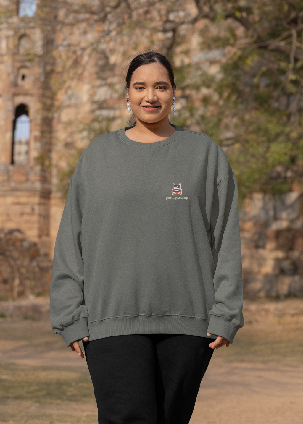 Edge of Evening Garment-Dyed Crewneck in Pepper, worn by a woman by an old stone building, with the Edge of Evening logo and portage.camp on the chest. Digital product mockup.