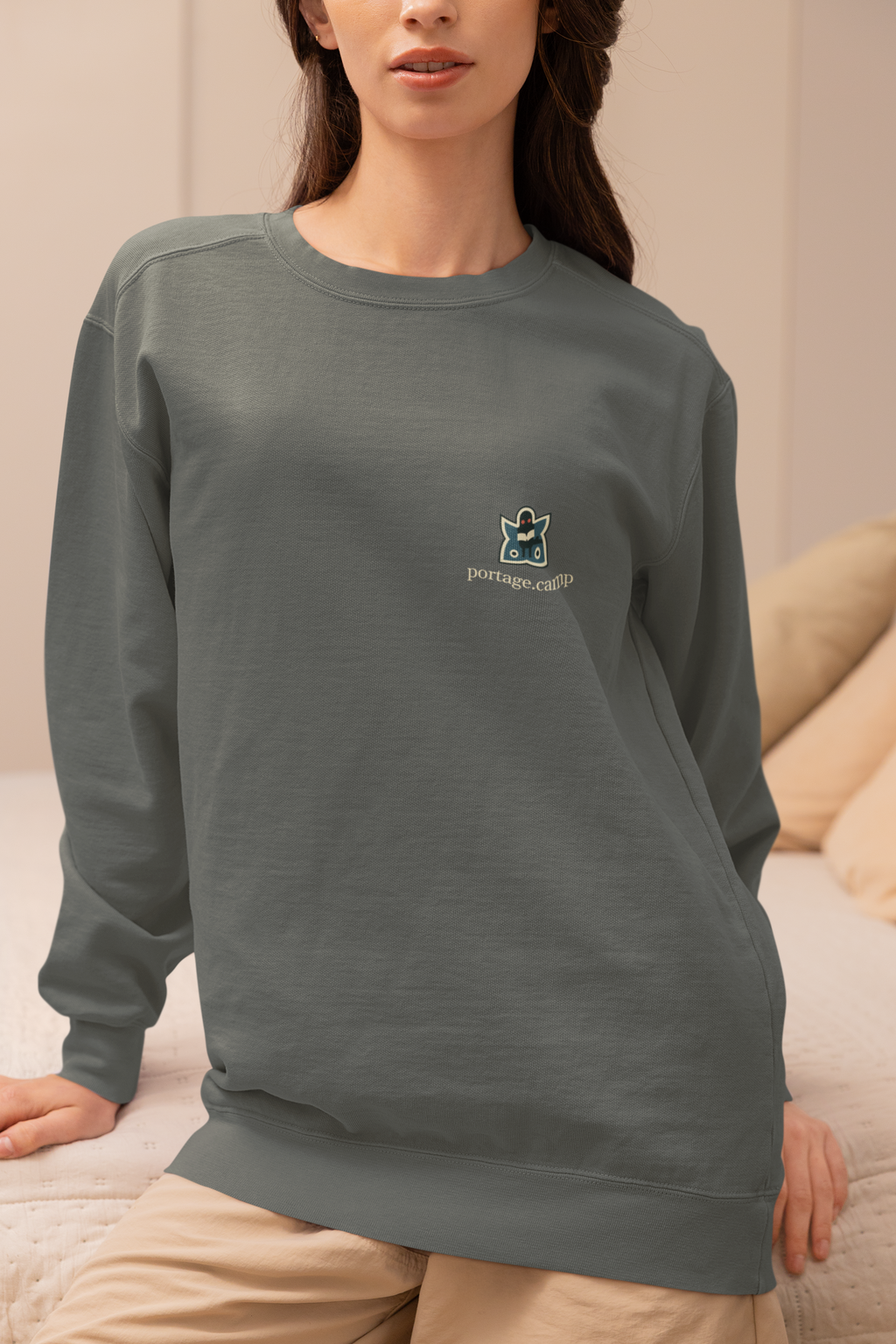 Night Library Garment-Dyed Crewneck in Pepper, worn by a woman seated on a bed at home, with the Night Library logo and portage.camp on the chest. Digital product mockup.