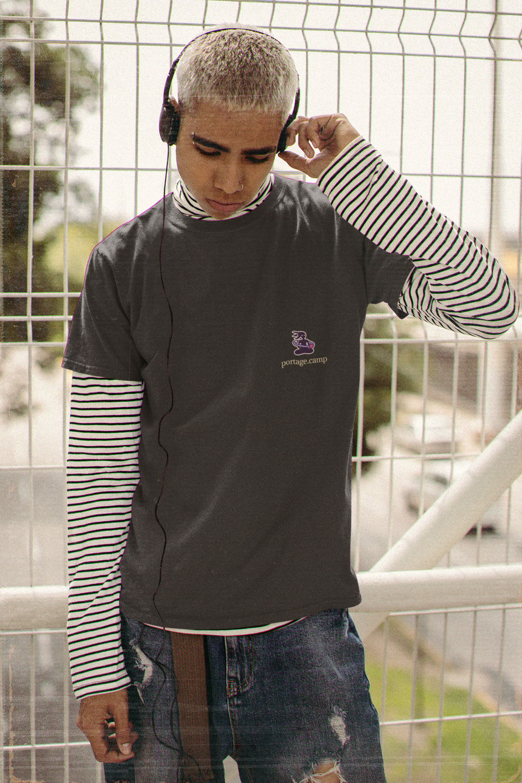 Orbit Keeper Garment-Dyed Tee in Black, worn by a man in headphones by a fence, with the Orbit Keeper logo and portage.camp on the chest. Digital product mockup.