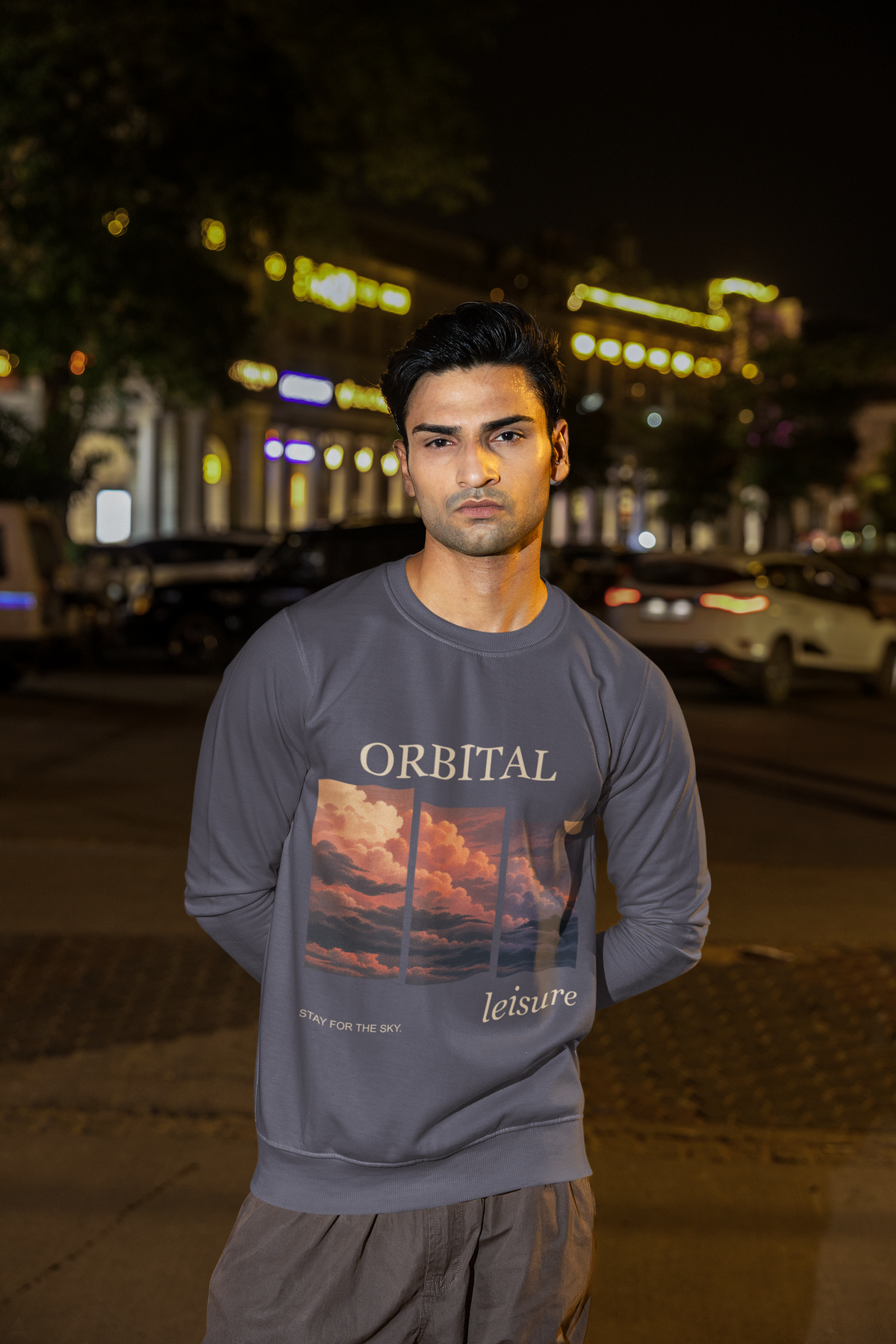 Stay for the Sky Garment-Dyed Crewneck (Front Print) in Denim, worn by a man on a city street at night, showing the large print on the front. Digital product mockup.