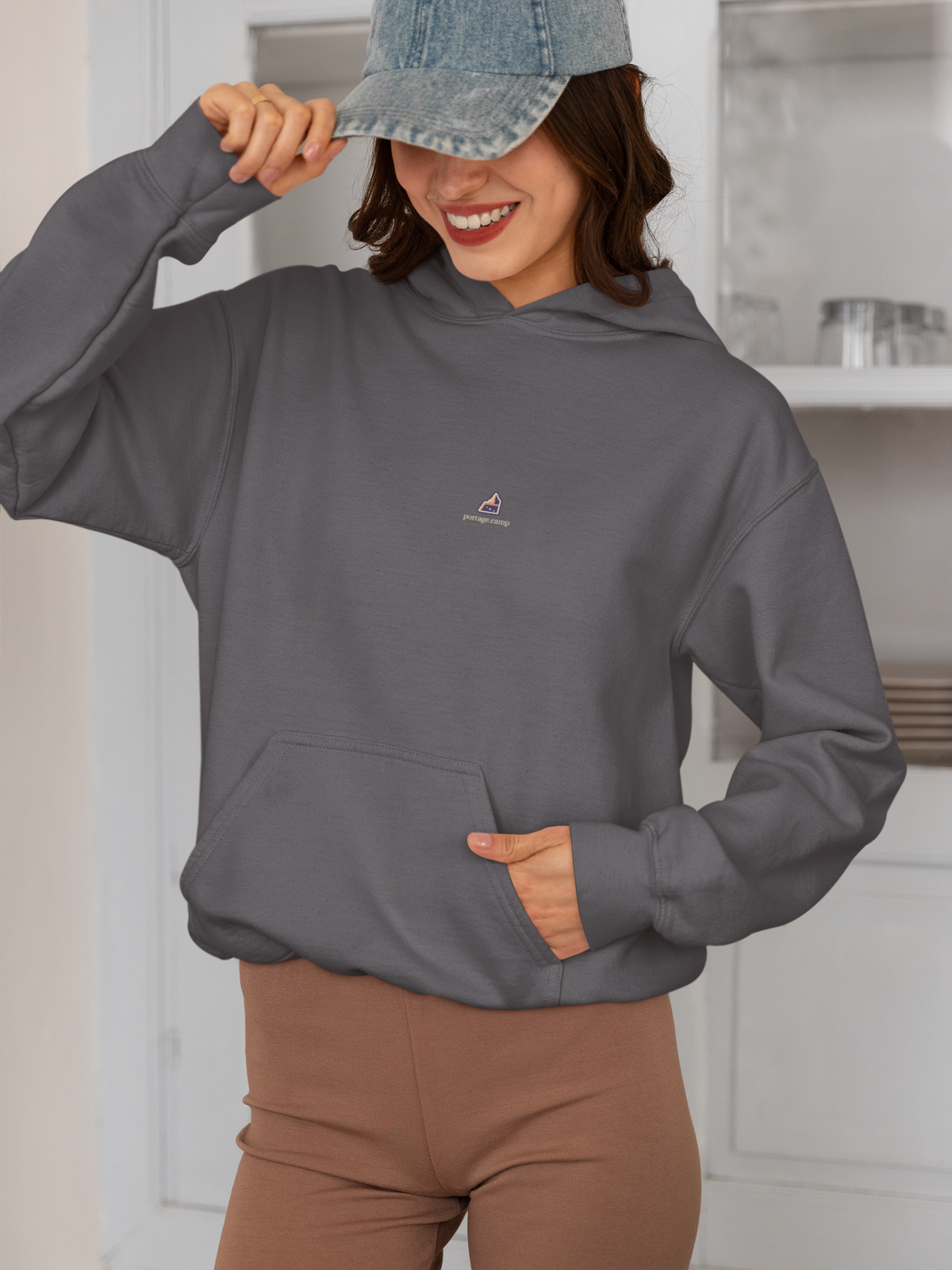 A Different Sky Midweight Hoodie in Charcoal, worn by a woman smiling in a kitchen, with the A Different Sky logo and portage.camp on the chest. Digital product mockup.