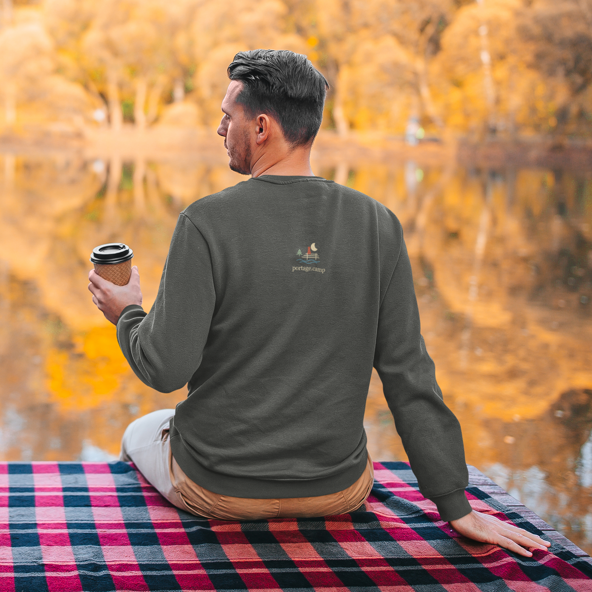 Quiet Hours Garment-Dyed Crewneck (Front Print) in Pepper, worn by a man seen from behind seated by a lake in autumn, showing the portage.camp emblem at the upper back. Digital product mockup.
