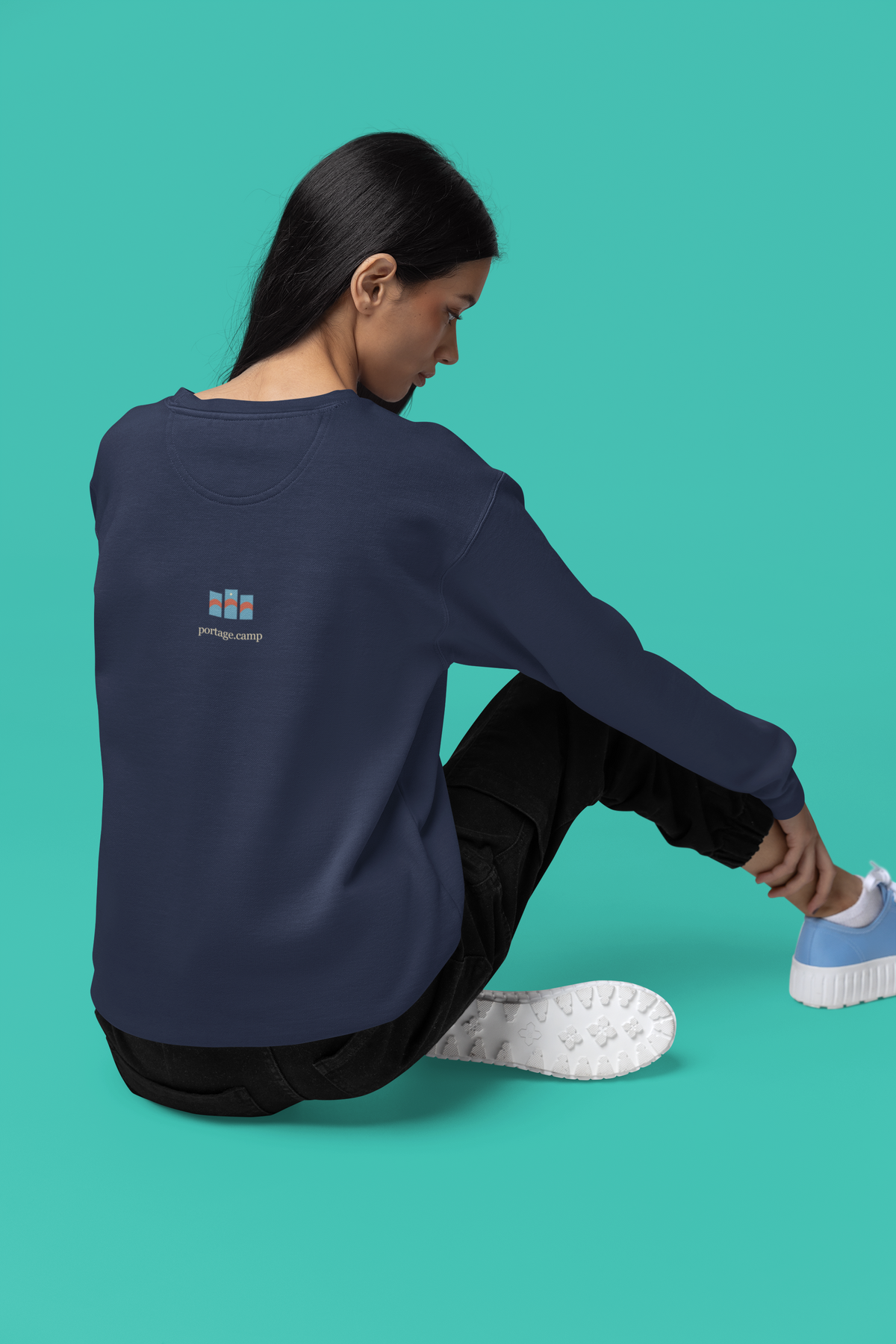 Stay for the Sky Garment-Dyed Crewneck (Front Print) in True Navy, worn by a woman seen from behind seated on a studio floor, showing the portage.camp emblem at the upper back. Digital product mockup.