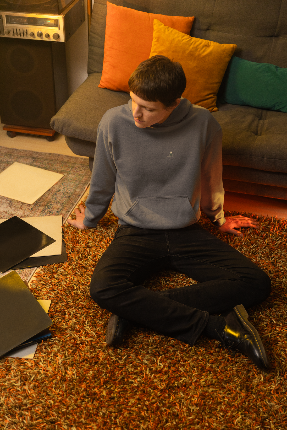 Small Hours Midweight Hoodie in Charcoal, worn by a man seated on the floor among records, with the Small Hours logo and portage.camp on the chest. Digital product mockup.