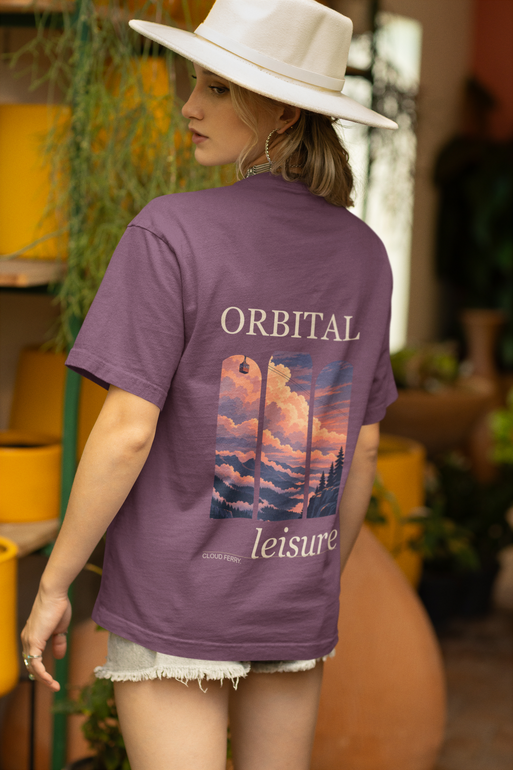 Cloud Ferry Garment-Dyed Tee in Berry, worn by a woman seen from behind in a sun hat among potted plants, showing the large print on the back. Digital product mockup.