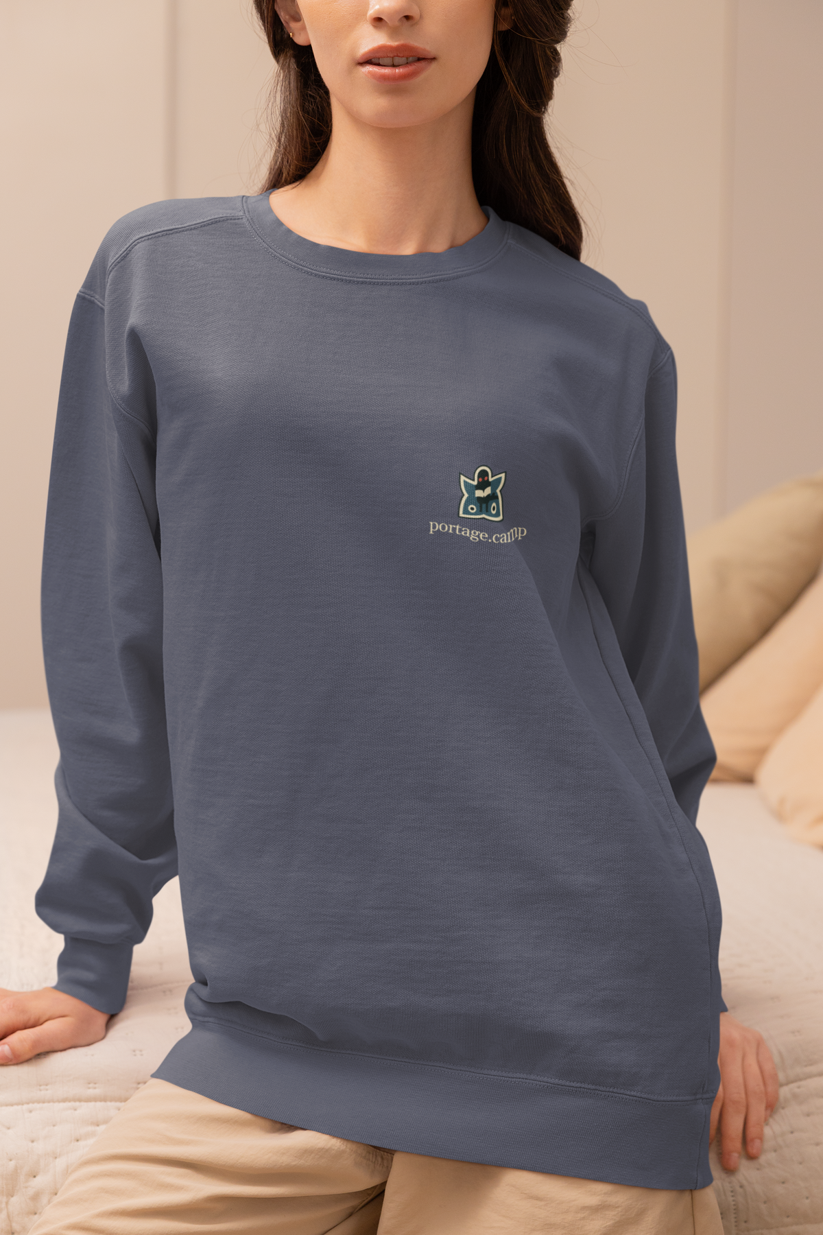 Night Library Garment-Dyed Crewneck in Denim, worn by a woman seated on a bed at home, with the Night Library logo and portage.camp on the chest. Digital product mockup.