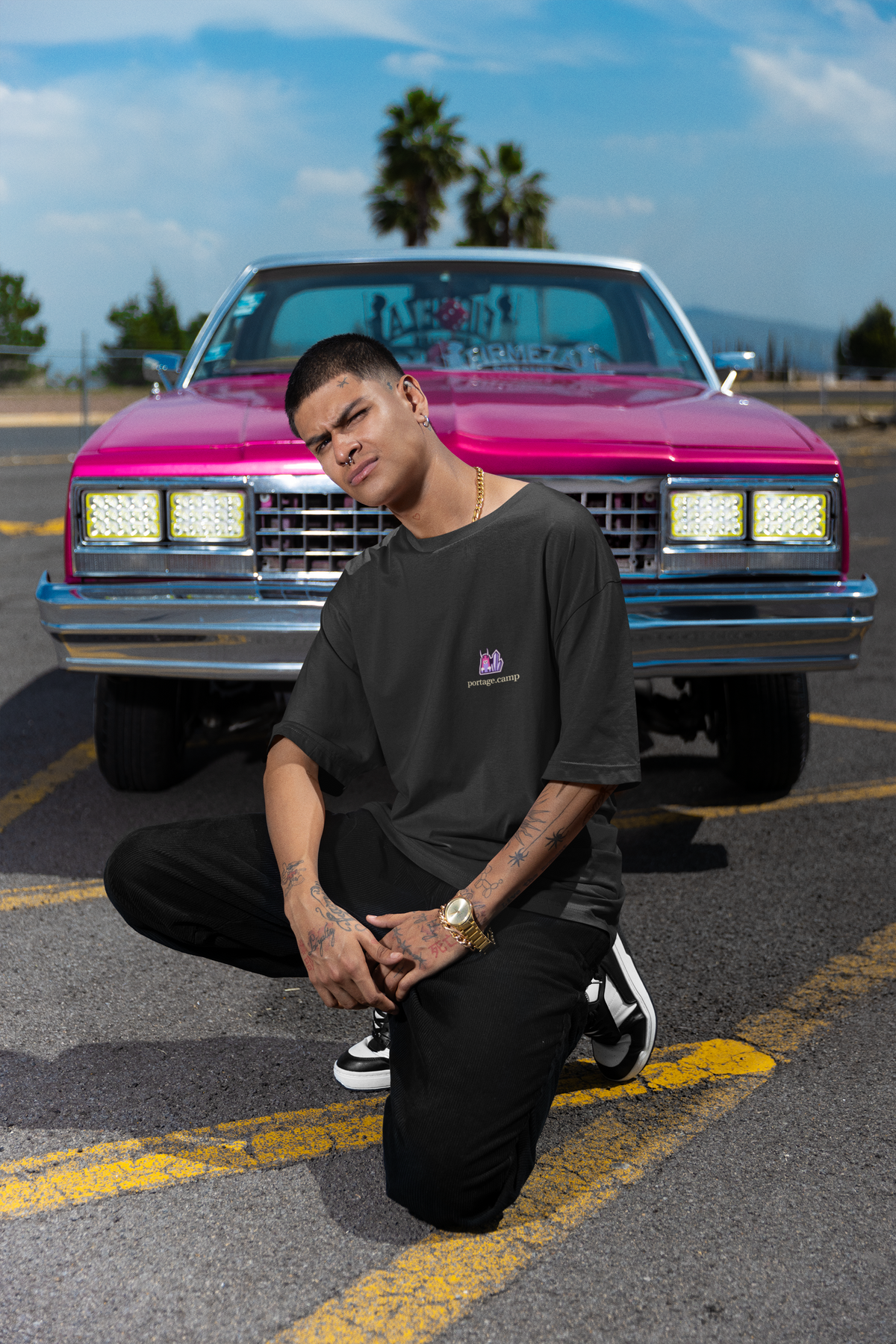 Amethyst After Hours Garment-Dyed Tee in Black, worn by a man crouched in front of a pink classic car, with the Amethyst After Hours logo and portage.camp on the chest. Digital product mockup.
