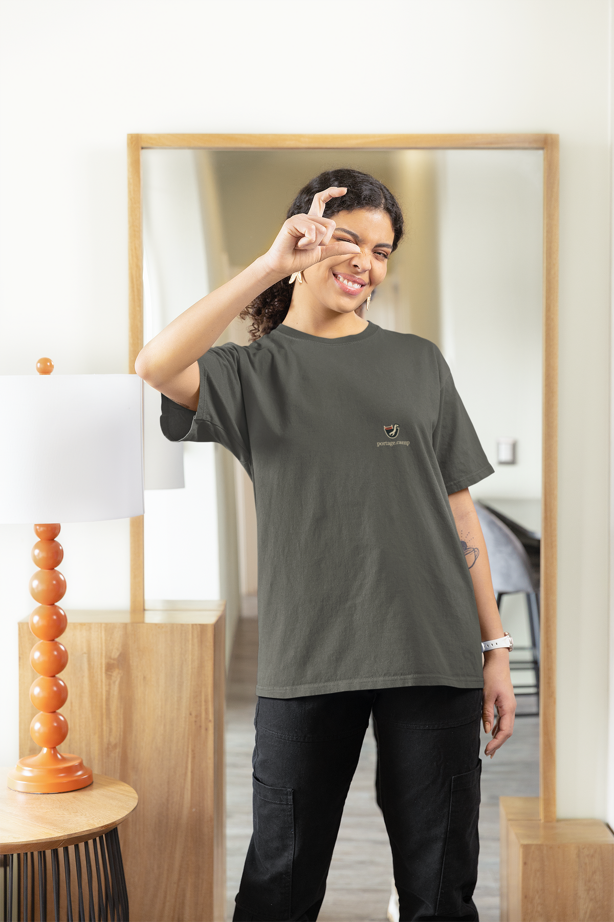 Seen Briefly Garment-Dyed Tee in Pepper, worn by a woman at home, pretending to take a photo, with the Seen Briefly logo and portage.camp on the chest. Digital product mockup.
