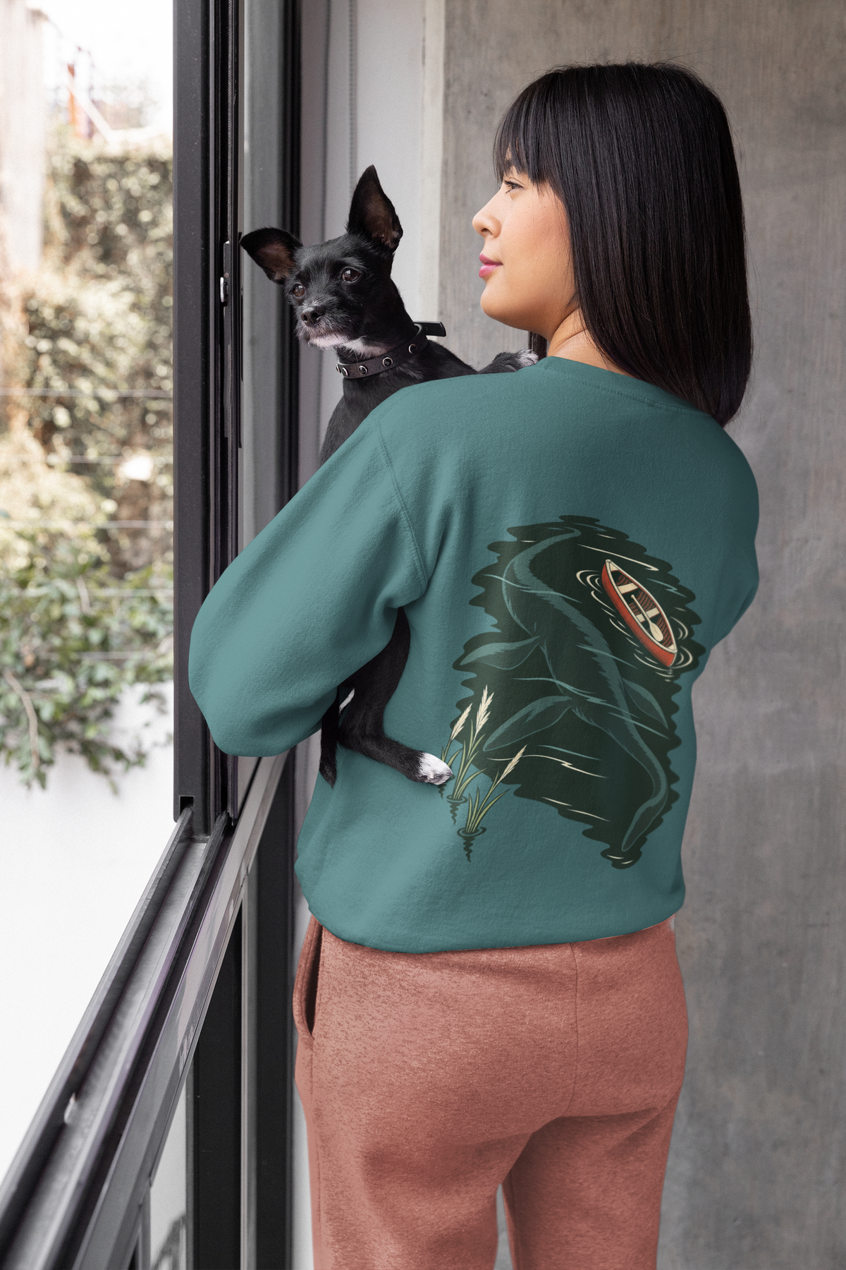 Seen Briefly Garment-Dyed Crewneck in Blue Spruce, worn by a woman seen from behind at home, holding her dog, showing the large print on the back. Digital product mockup.