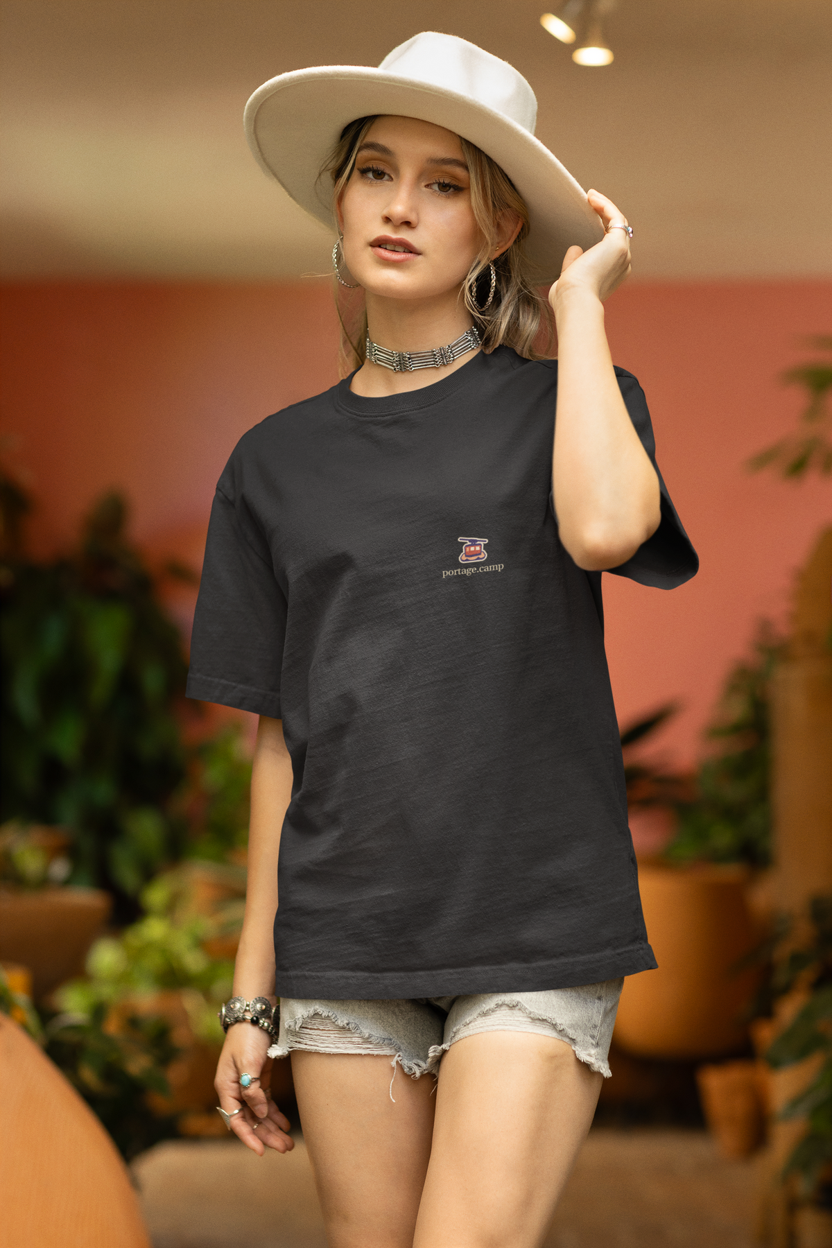 Cloud Ferry Garment-Dyed Tee in Black, worn by a woman in a sun hat among potted plants, with the Cloud Ferry logo and portage.camp on the chest. Digital product mockup.