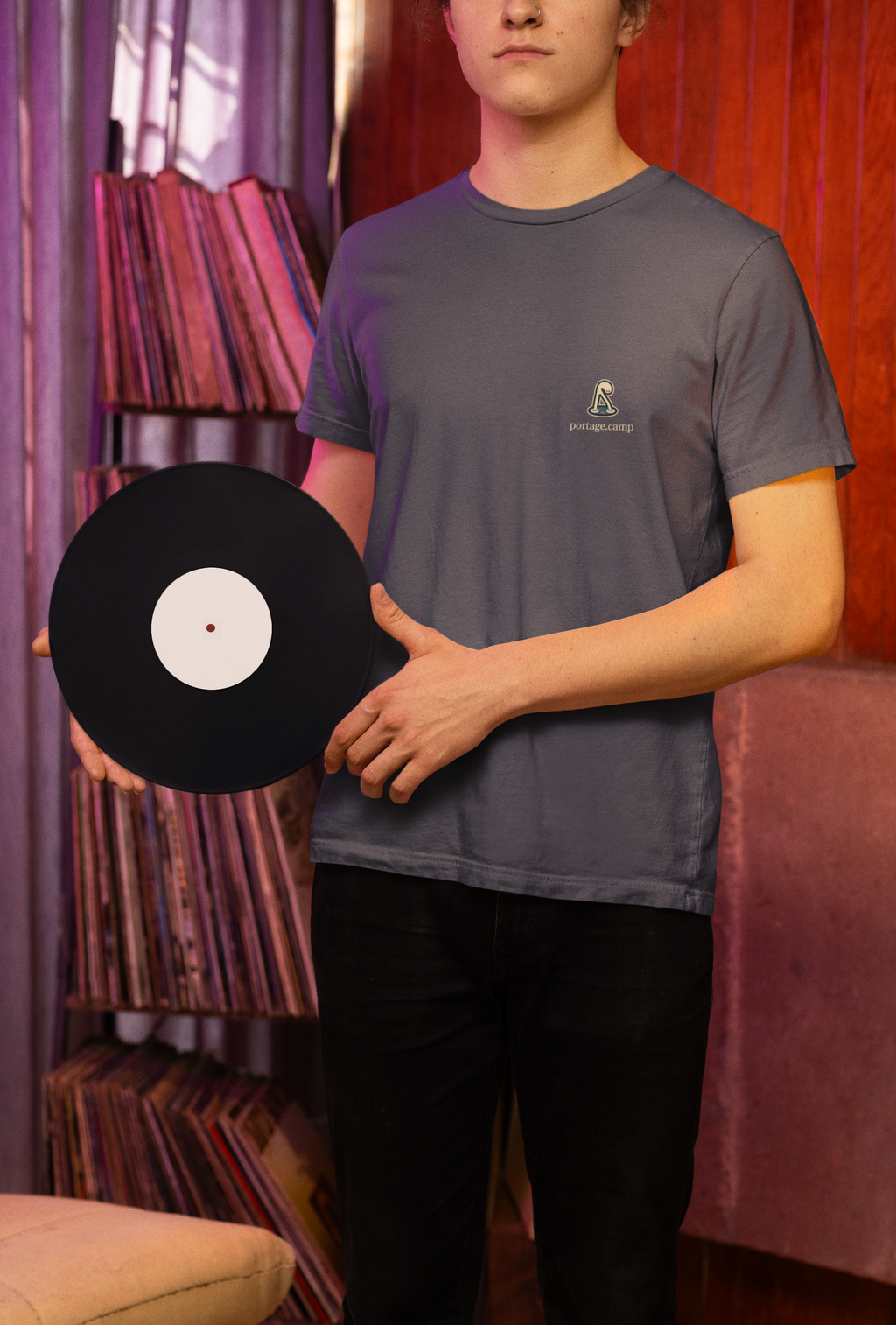 Small Hours Garment-Dyed Tee in Navy, worn by a man holding a vinyl record in a record shop, with the Small Hours logo and portage.camp on the chest. Digital product mockup.