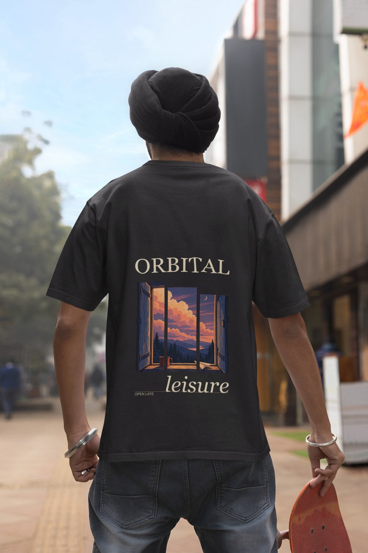 Open Late Garment-Dyed Tee in Black, worn by a man seen from behind in a turban on a busy street, showing the large print on the back. Digital product mockup.