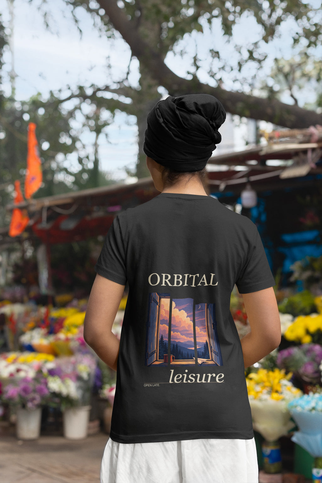 Open Late Garment-Dyed Tee in Black, worn by a woman seen from behind in a turban by a flower market, showing the large print on the back. Digital product mockup.