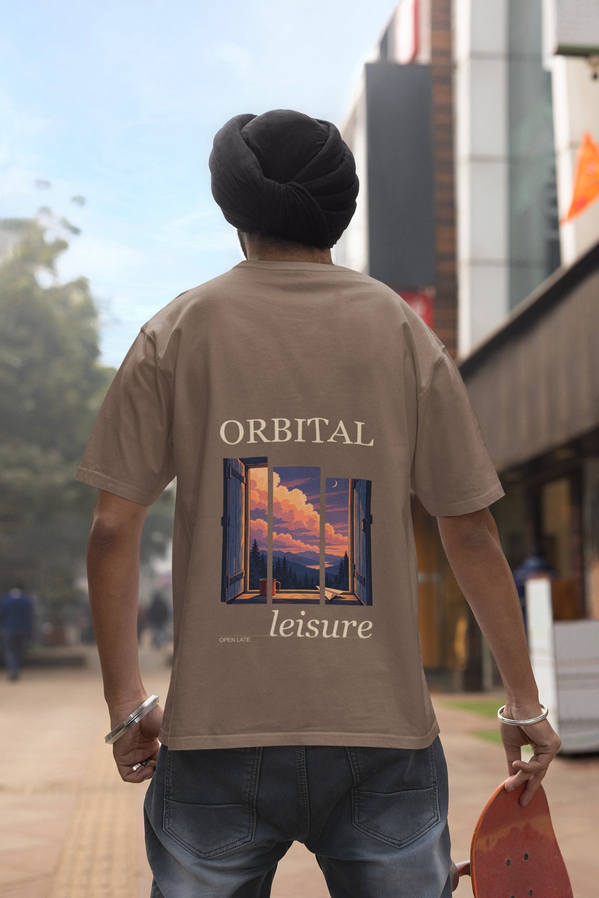 Open Late Garment-Dyed Tee in Espresso, worn by a man seen from behind in a turban on a busy street, showing the large print on the back. Digital product mockup.