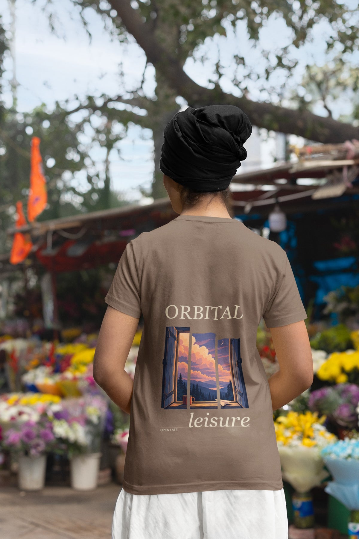 Open Late Garment-Dyed Tee in Espresso, worn by a woman seen from behind in a turban by a flower market, showing the large print on the back. Digital product mockup.