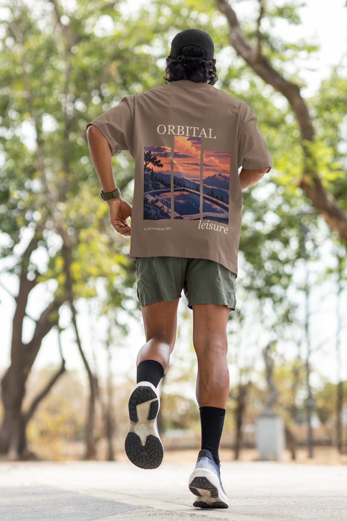 A Different Sky Garment-Dyed Tee in Espresso, worn by a man seen from behind jogging through a park, showing the large print on the back. Digital product mockup.