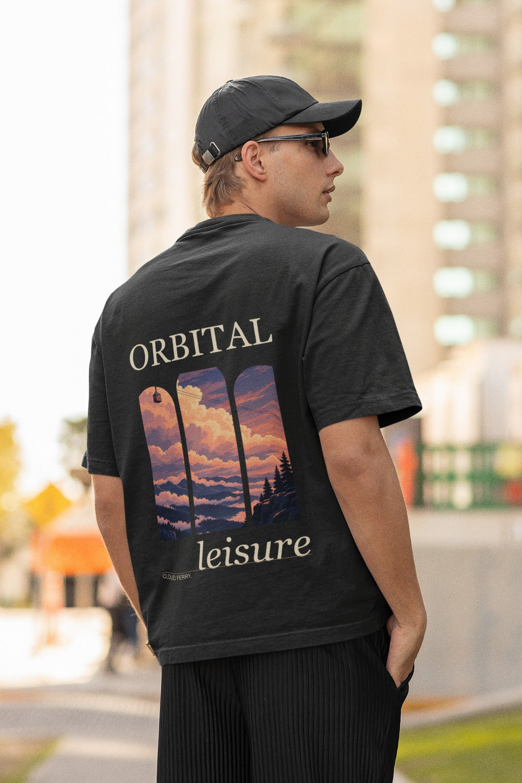 Cloud Ferry Garment-Dyed Tee in Black, worn by a man seen from behind in a cap and sunglasses on a city street, showing the large print on the back. Digital product mockup.