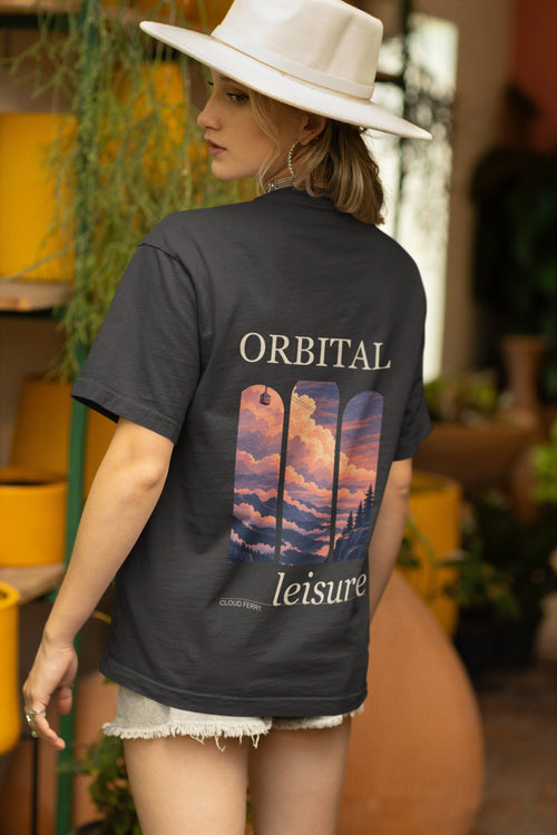 Cloud Ferry Garment-Dyed Tee in Black, worn by a woman seen from behind in a sun hat among potted plants, showing the large print on the back. Digital product mockup.