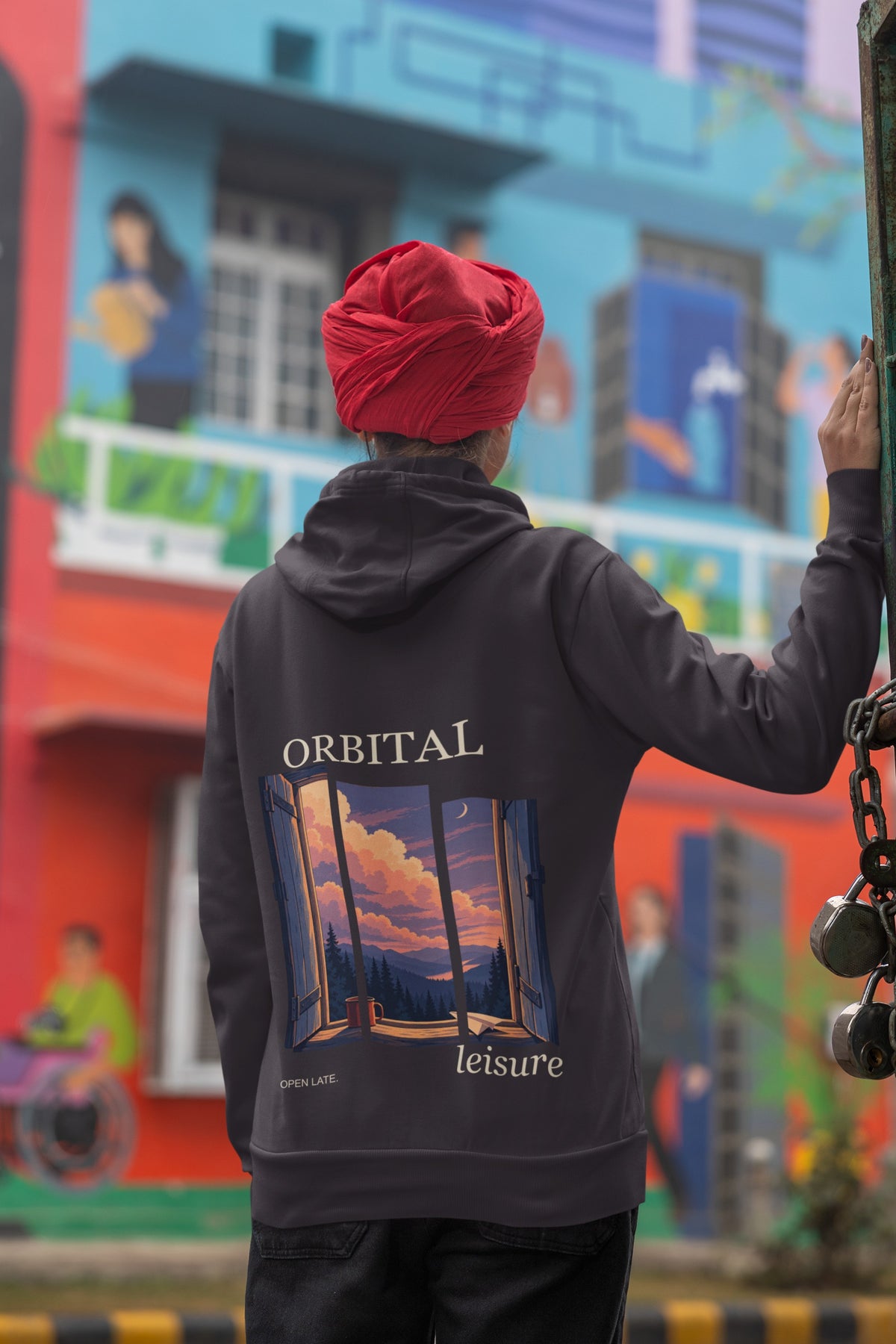 Open Late Midweight Hoodie in Black, worn by a woman seen from behind in a turban by a painted mural, showing the large print on the back. Digital product mockup.