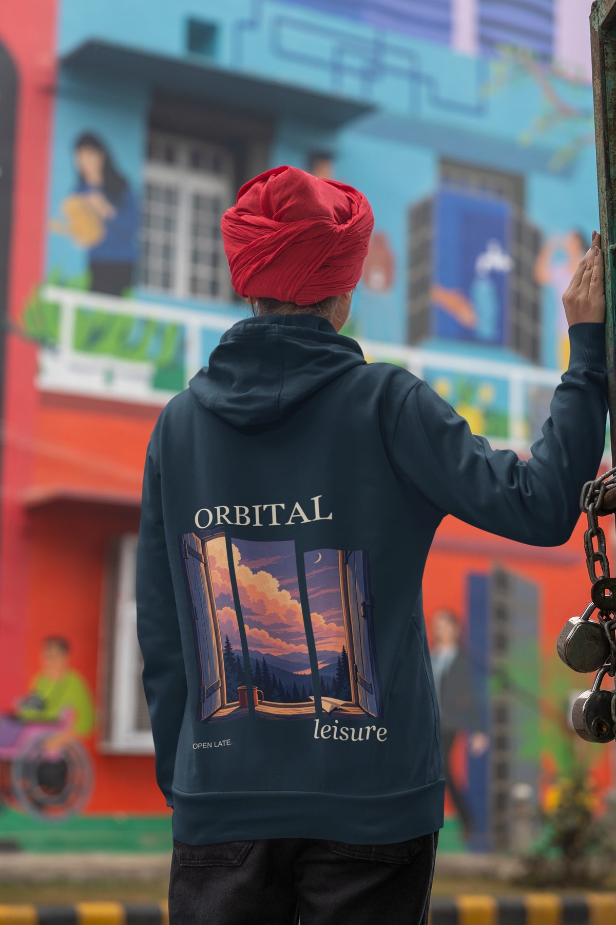 Open Late Midweight Hoodie in Classic Navy, worn by a woman seen from behind in a turban by a painted mural, showing the large print on the back. Digital product mockup.