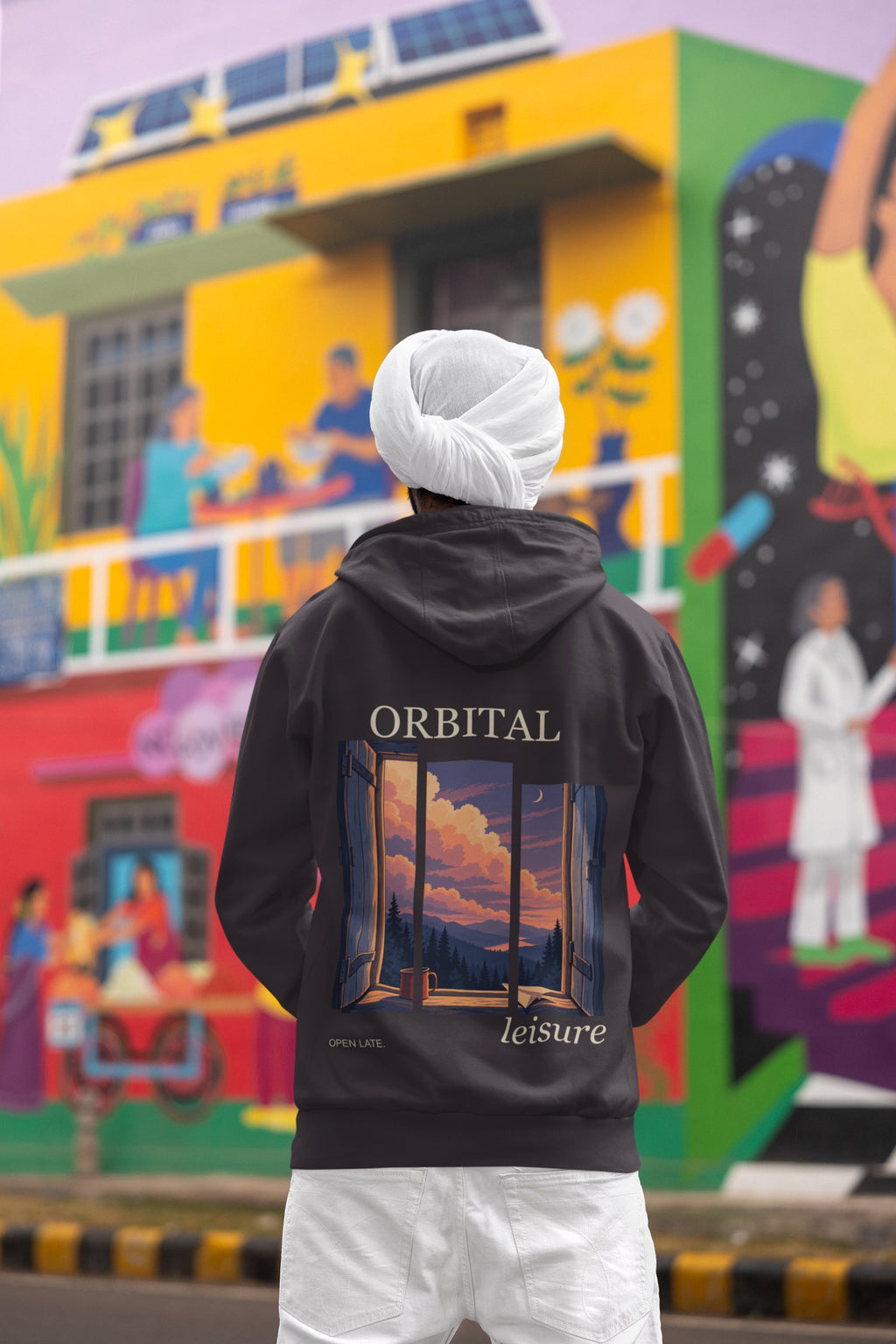 Open Late Midweight Hoodie in Black, worn by a man seen from behind in a turban by a painted mural, showing the large print on the back. Digital product mockup.