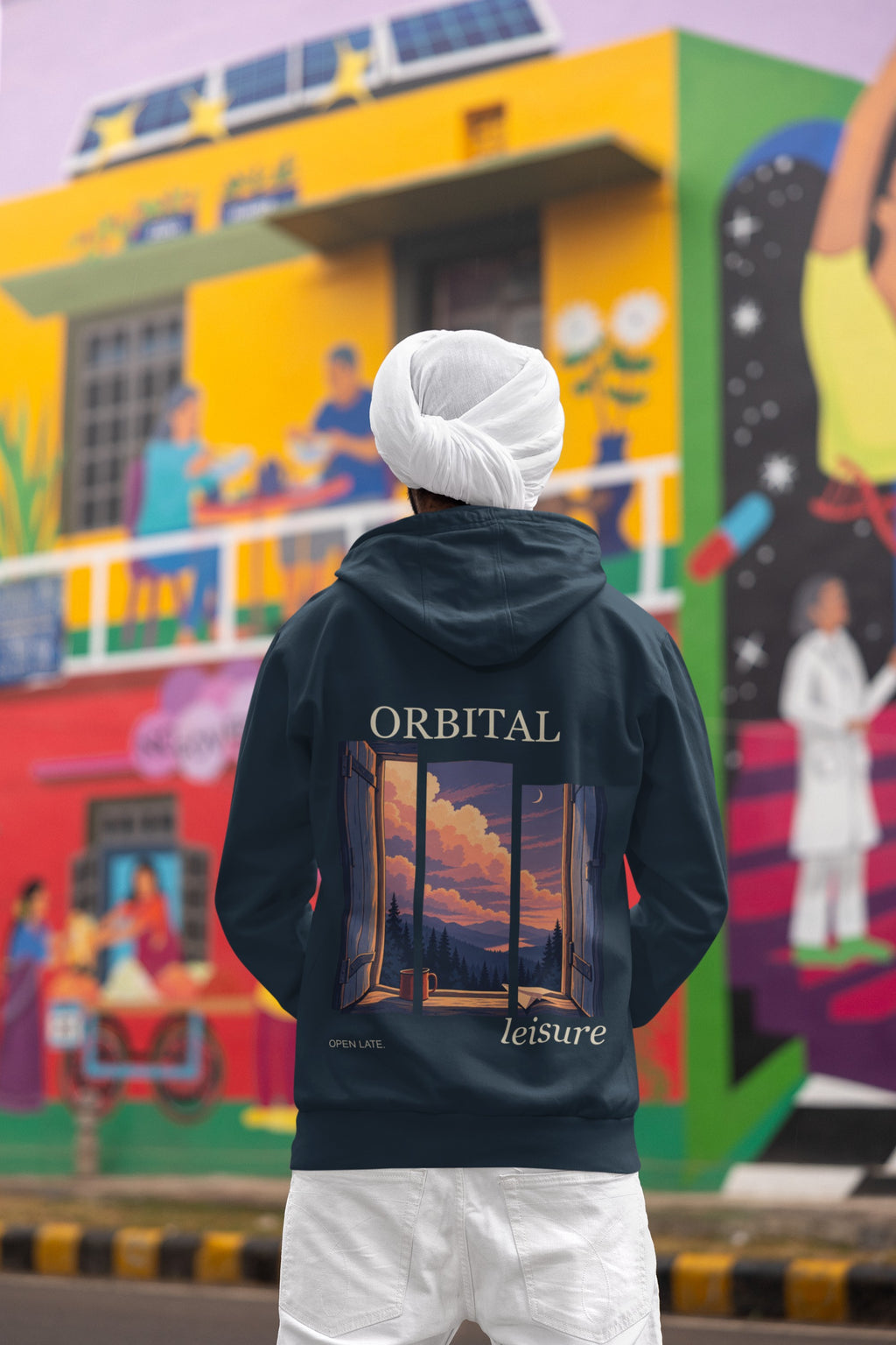 Open Late Midweight Hoodie in Classic Navy, worn by a man seen from behind in a turban by a painted mural, showing the large print on the back. Digital product mockup.