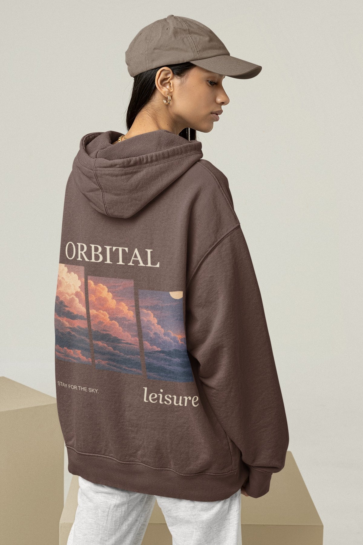 Stay for the Sky Heavyweight Hoodie in Brown, worn by a woman seen from behind in a studio, showing the large print on the back. Digital product mockup.