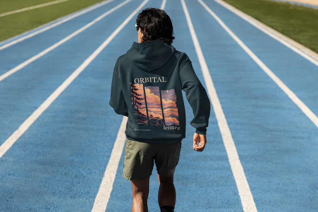 Edge of Evening Midweight Hoodie in Classic Navy, worn by a man seen from behind on a running track, showing the large print on the back. Digital product mockup.