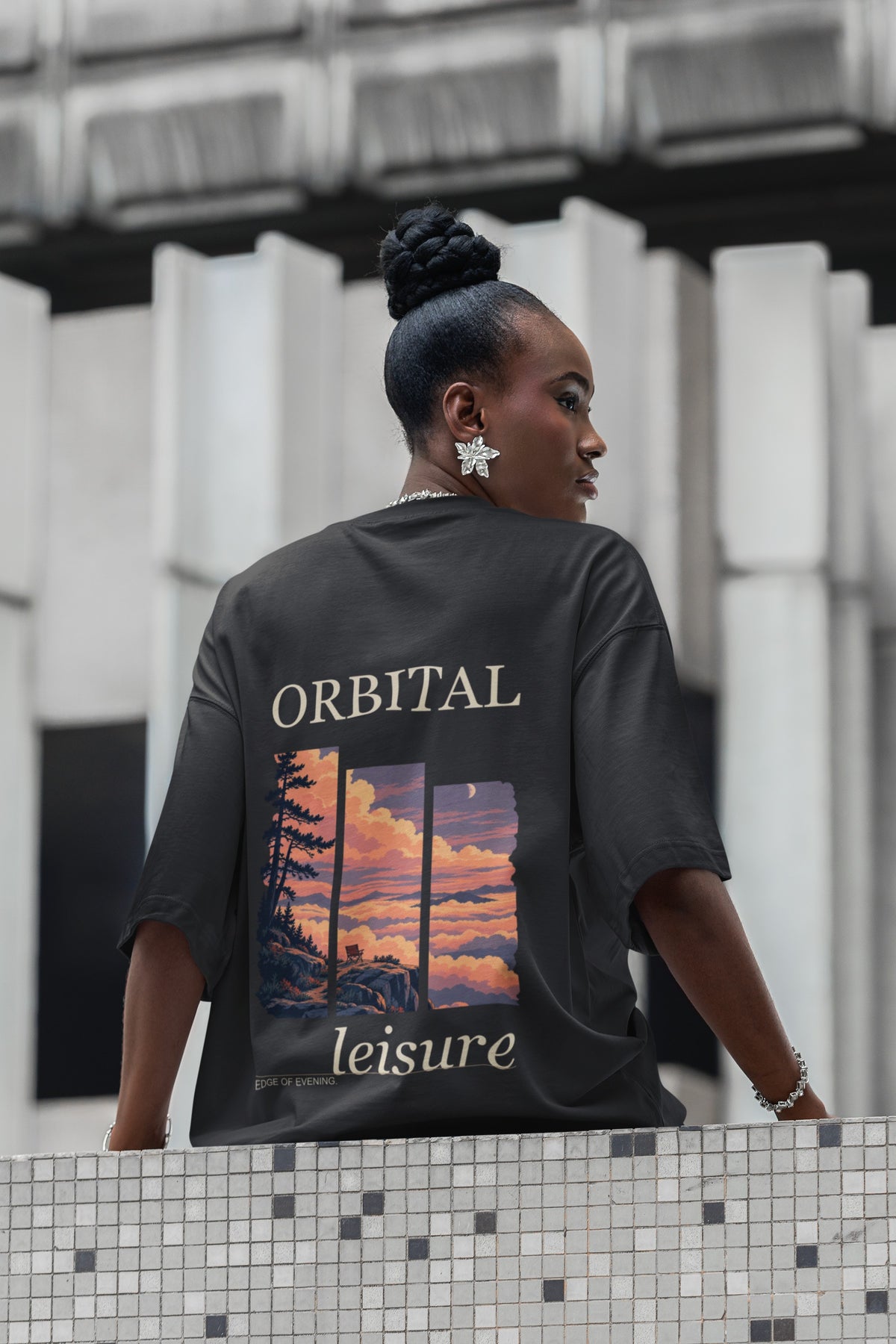 Edge of Evening Garment-Dyed Tee in Black, worn by a woman seen from behind on a city street lined with columns, showing the large print on the back. Digital product mockup.
