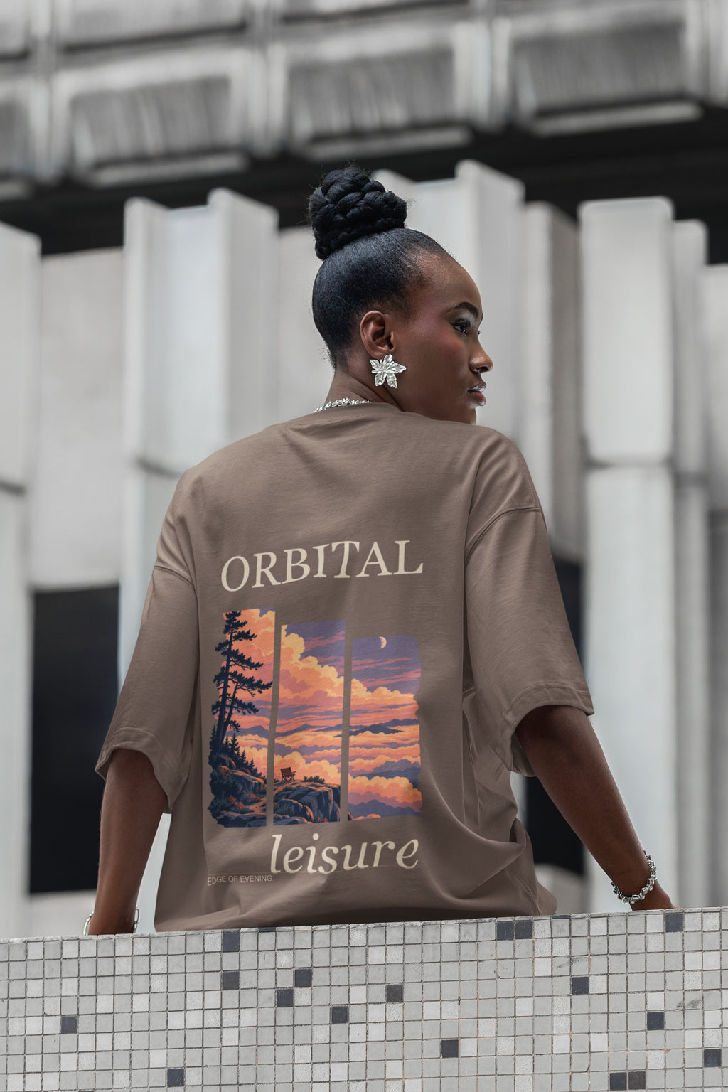 Edge of Evening Garment-Dyed Tee in Espresso, worn by a woman seen from behind on a city street lined with columns, showing the large print on the back. Digital product mockup.