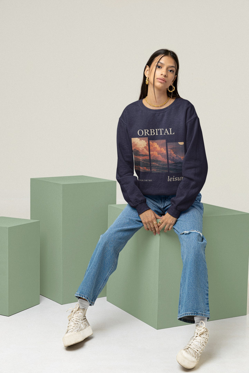 Stay for the Sky Garment-Dyed Crewneck (Front Print) in True Navy, worn by a woman seated on green studio blocks, showing the large print on the front. Digital product mockup.