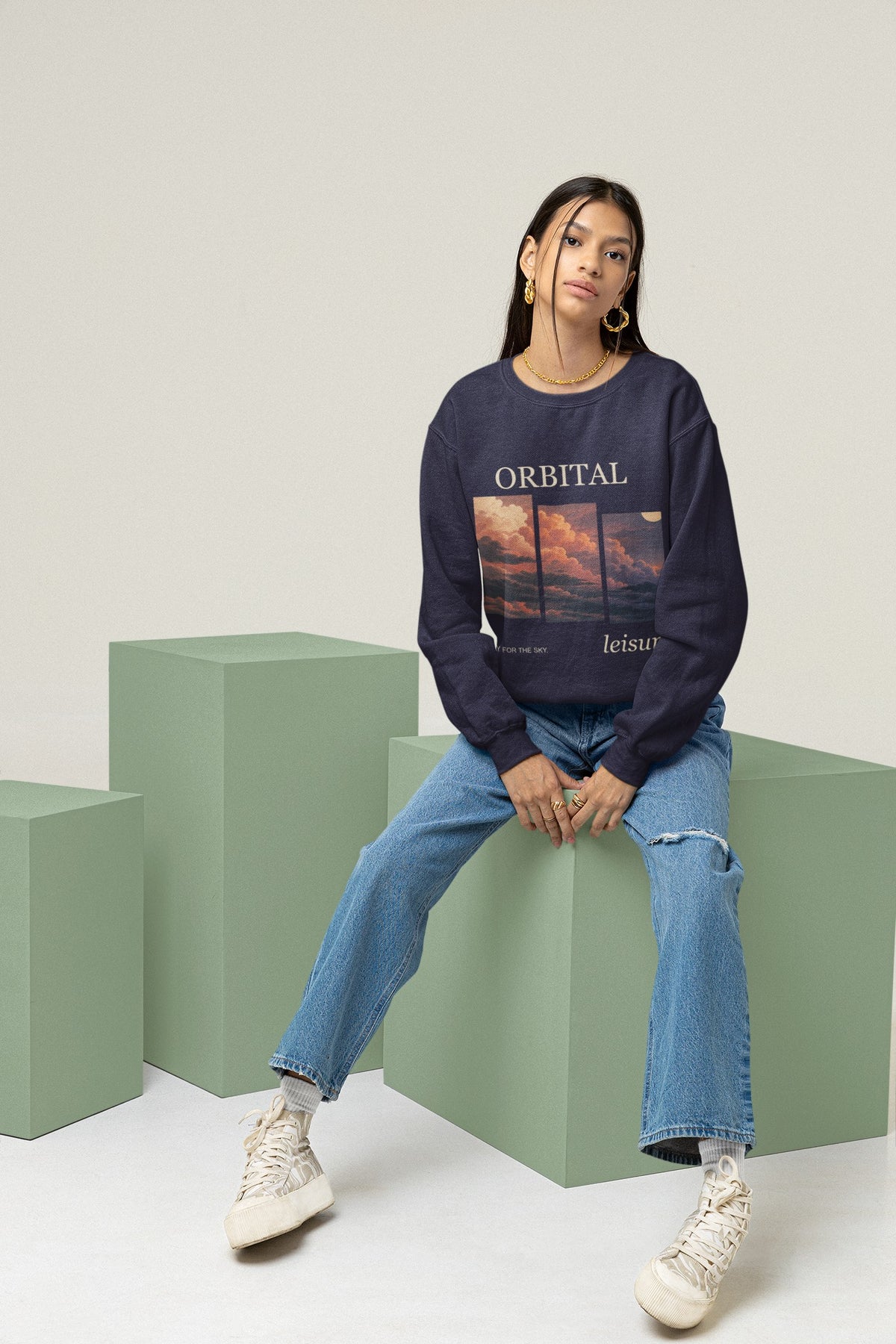 Stay for the Sky Garment-Dyed Crewneck (Front Print) in True Navy, worn by a woman seated on green studio blocks, showing the large print on the front. Digital product mockup.