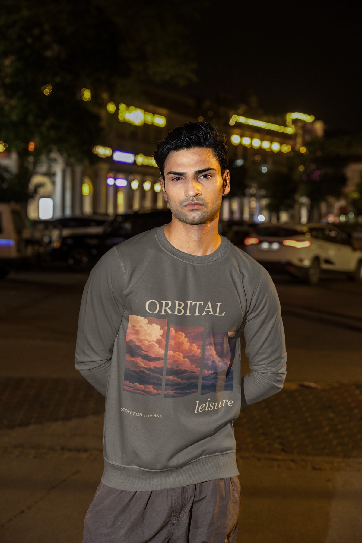 Stay for the Sky Garment-Dyed Crewneck (Front Print) in Pepper, worn by a man on a city street at night, showing the large print on the front. Digital product mockup.