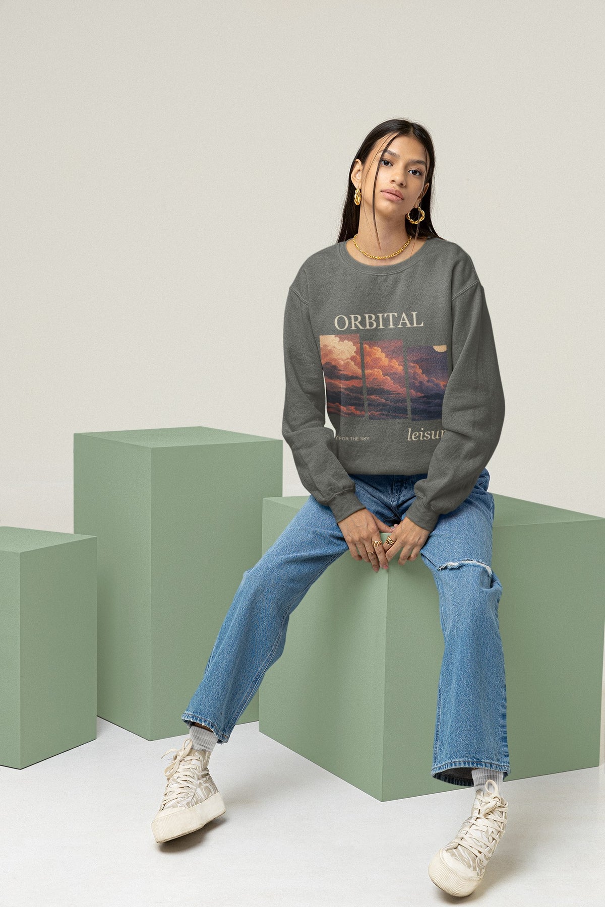 Stay for the Sky Garment-Dyed Crewneck (Front Print) in Pepper, worn by a woman seated on green studio blocks, showing the large print on the front. Digital product mockup.