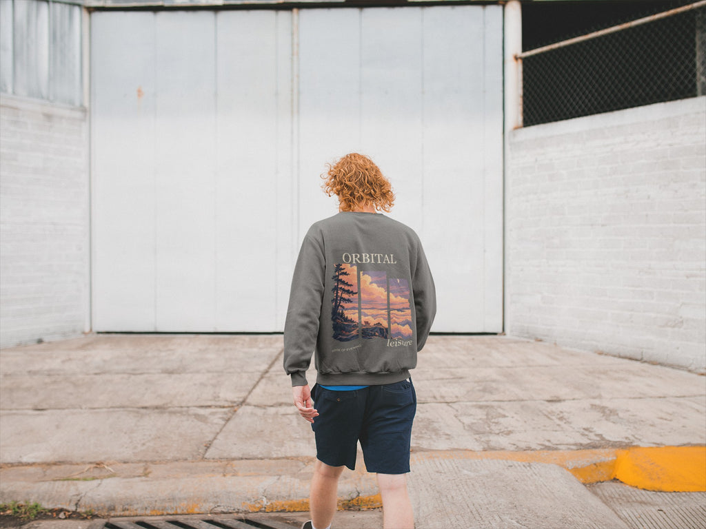 Edge of Evening Garment-Dyed Crewneck in Pepper, worn by a man seen from behind walking away across an empty yard, showing the large print on the back. Digital product mockup.