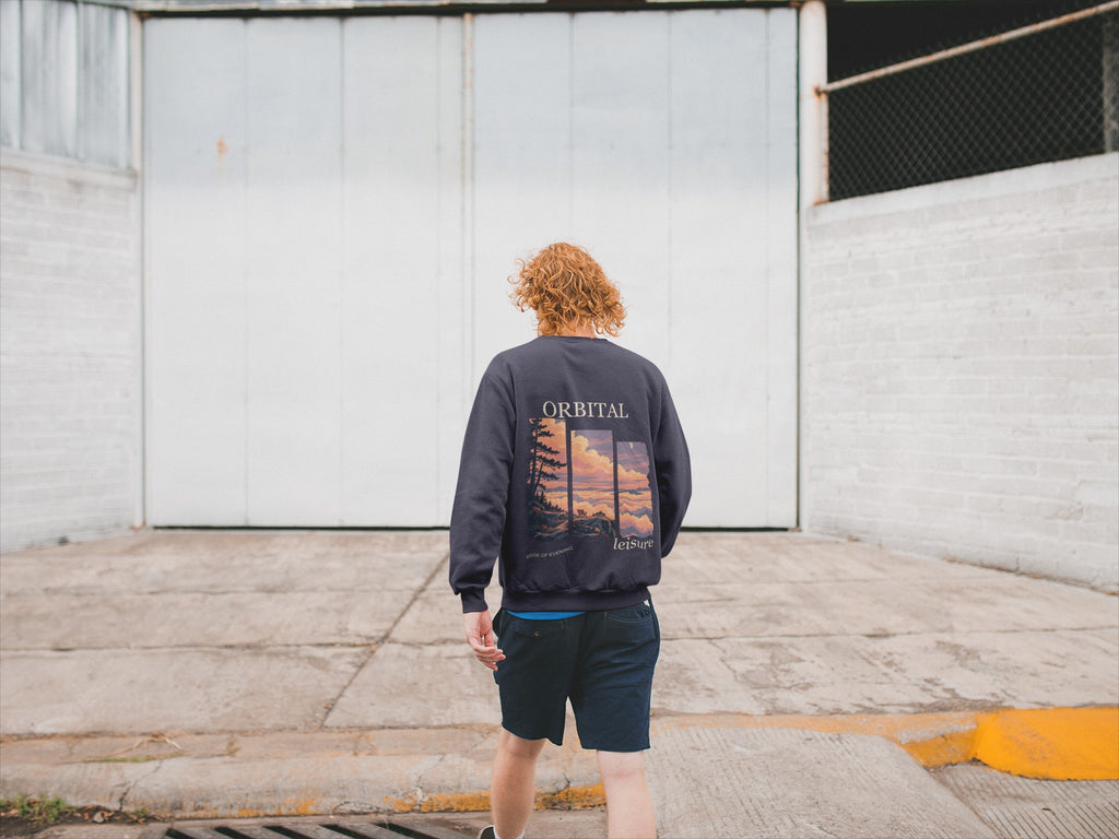 Edge of Evening Garment-Dyed Crewneck in True Navy, worn by a man seen from behind walking away across an empty yard, showing the large print on the back. Digital product mockup.