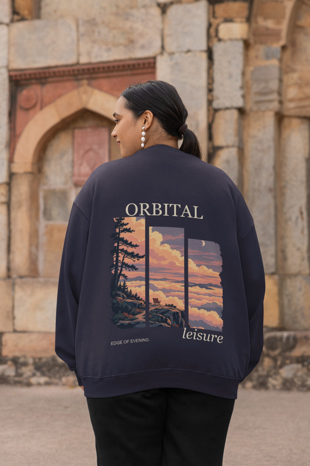 Edge of Evening Garment-Dyed Crewneck in True Navy, worn by a woman seen from behind by an old stone building, showing the large print on the back. Digital product mockup.
