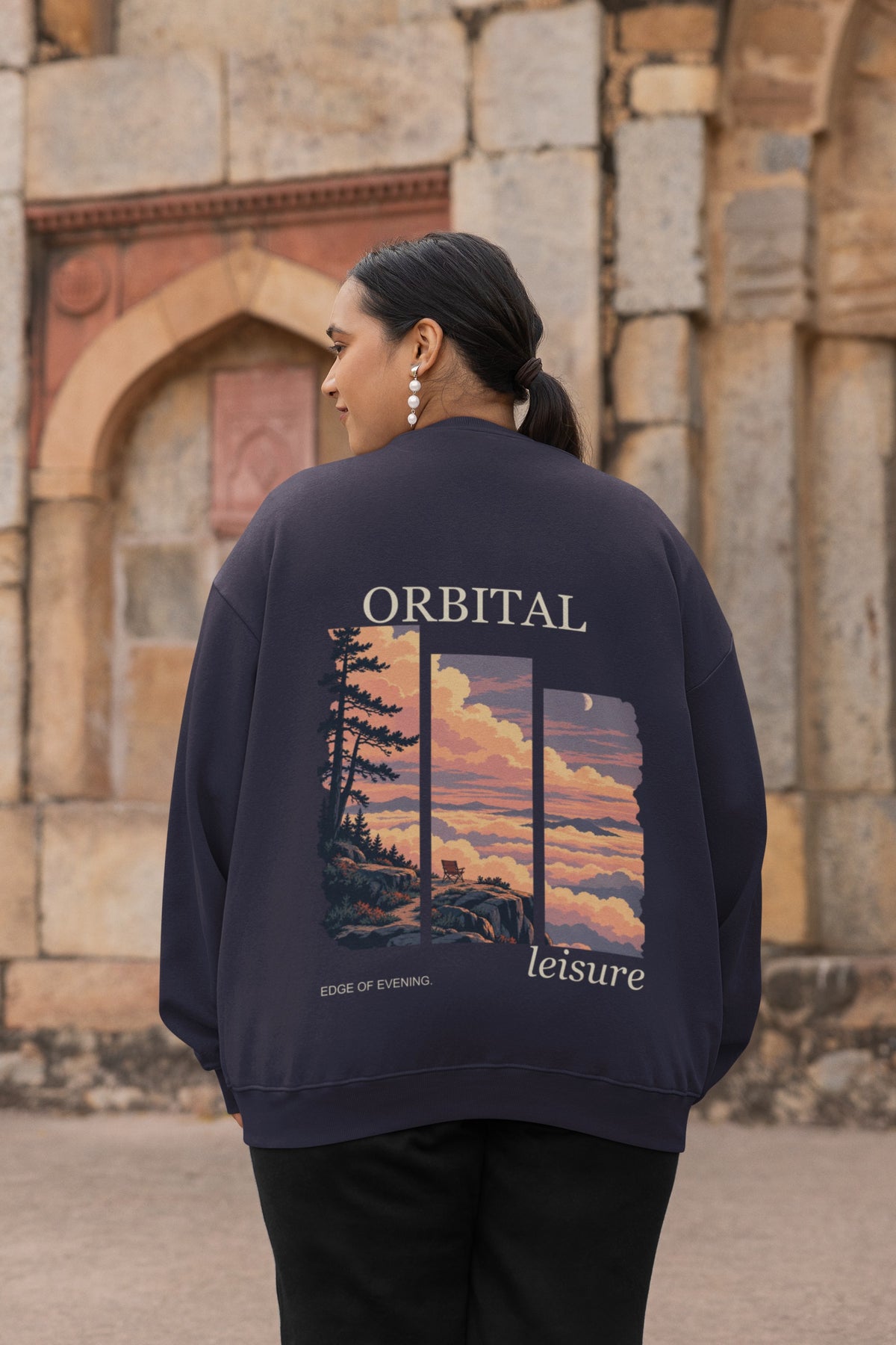 Edge of Evening Garment-Dyed Crewneck in True Navy, worn by a woman seen from behind by an old stone building, showing the large print on the back. Digital product mockup.