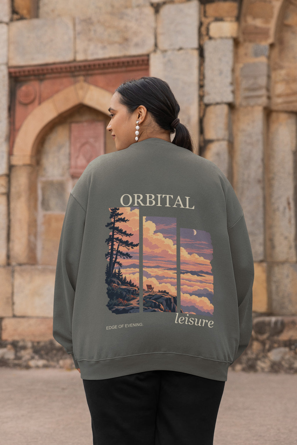 Edge of Evening Garment-Dyed Crewneck in Pepper, worn by a woman seen from behind by an old stone building, showing the large print on the back. Digital product mockup.