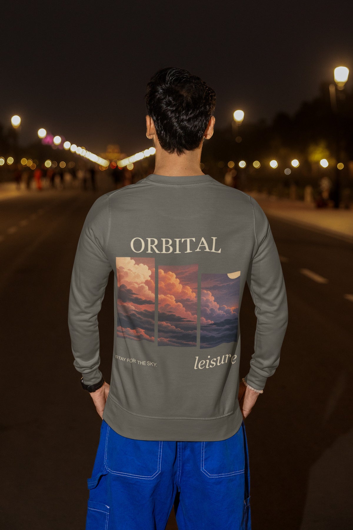 Stay for the Sky Garment-Dyed Crewneck in Pepper, worn by a man seen from behind on a city street at night, showing the large print on the back. Digital product mockup.