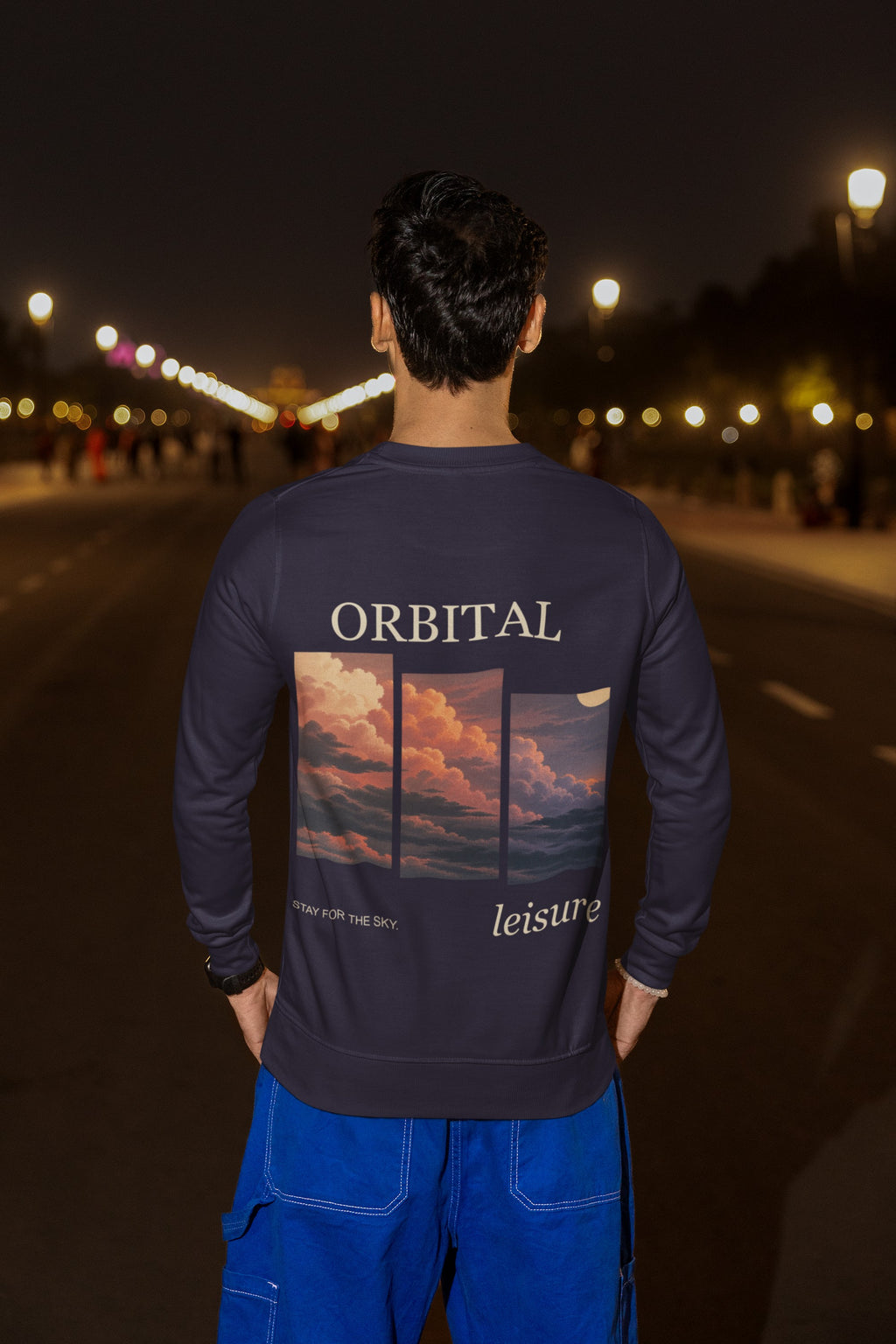 Stay for the Sky Garment-Dyed Crewneck in True Navy, worn by a man seen from behind on a city street at night, showing the large print on the back. Digital product mockup.