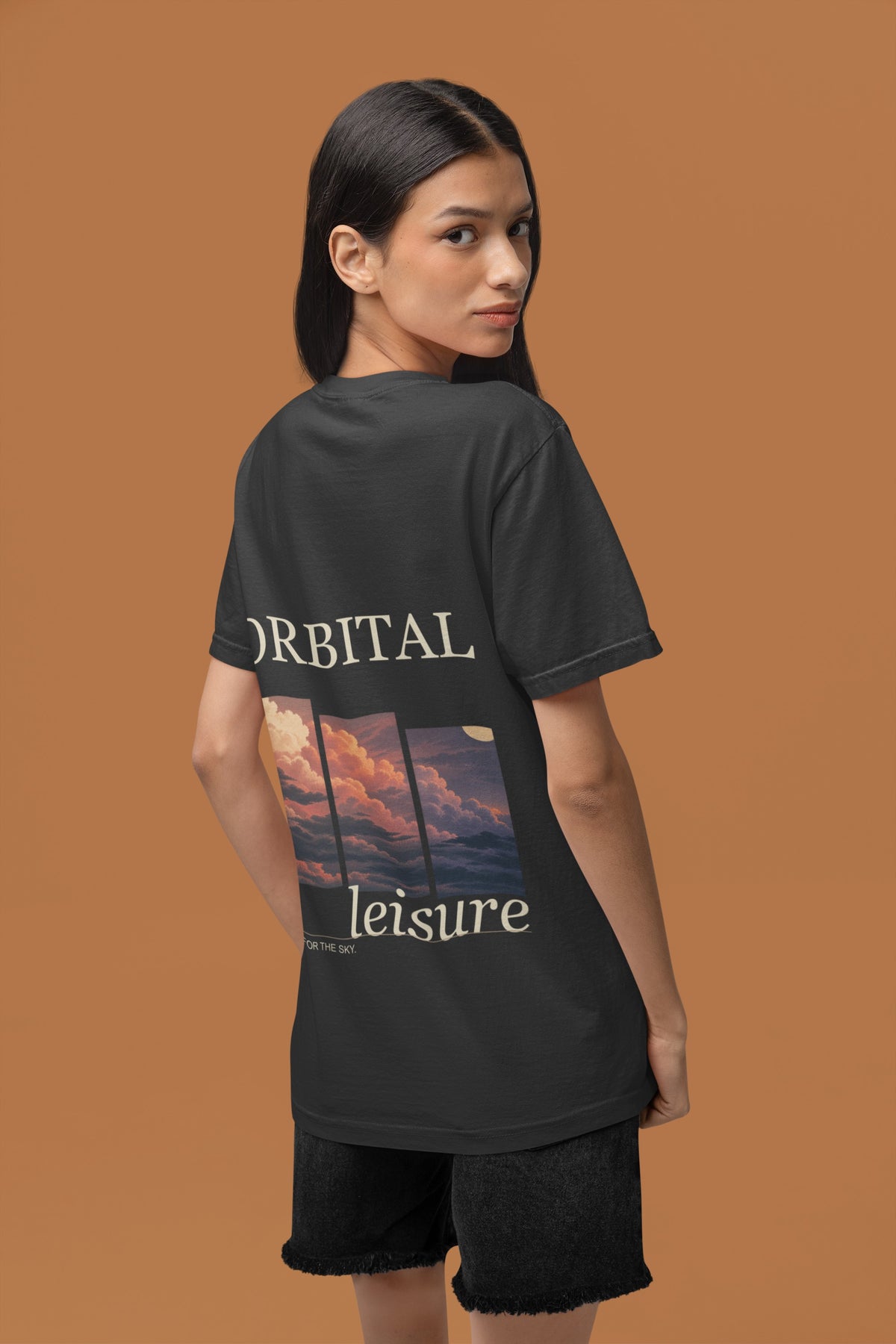 Stay for the Sky Garment-Dyed Tee in Black, worn by a woman seen from behind in a studio against a warm brown wall, showing the large print on the back. Digital product mockup.