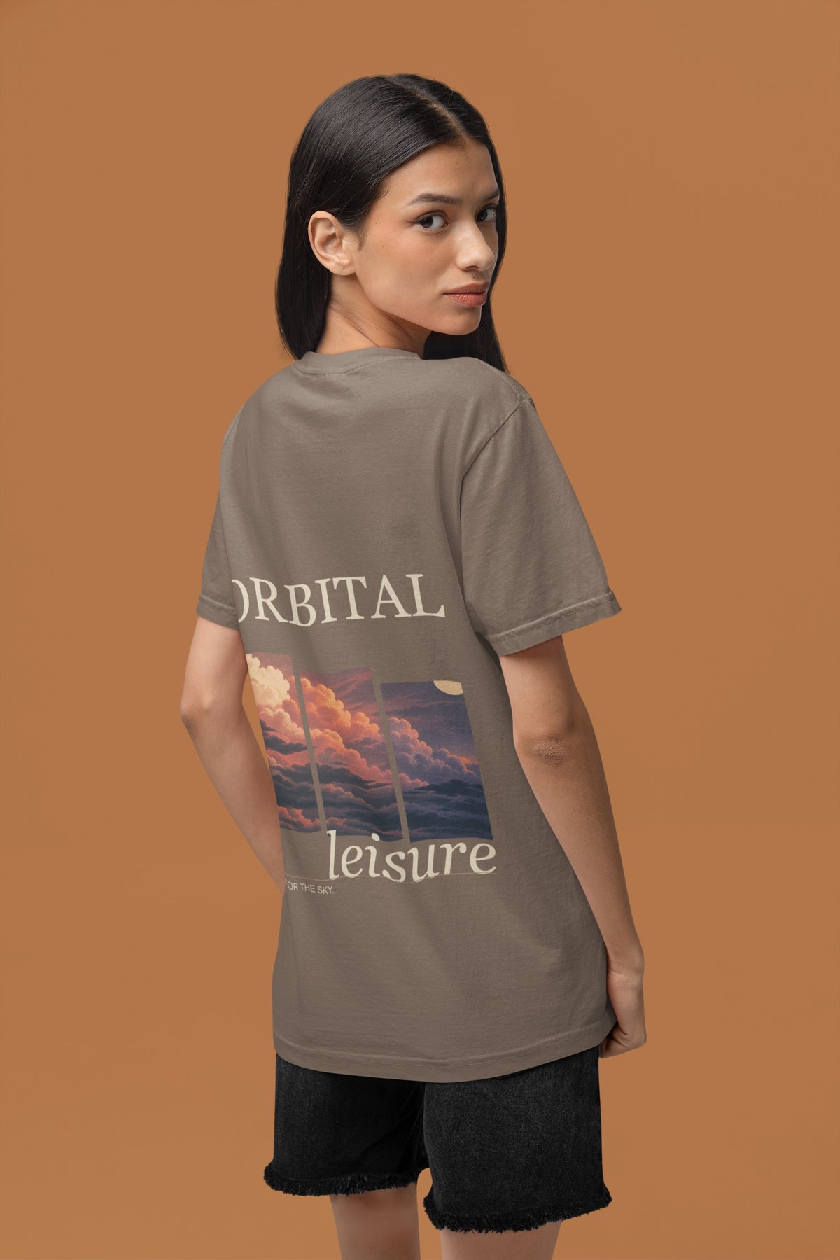 Stay for the Sky Garment-Dyed Tee in Espresso, worn by a woman seen from behind in a studio against a warm brown wall, showing the large print on the back. Digital product mockup.