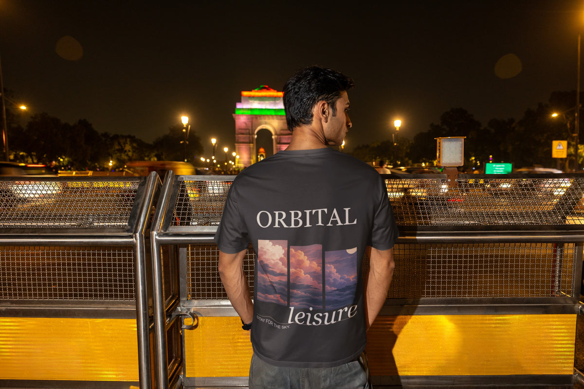 Stay for the Sky Garment-Dyed Tee in Black, worn by a man seen from behind on a bridge over the city at night, showing the large print on the back. Digital product mockup.